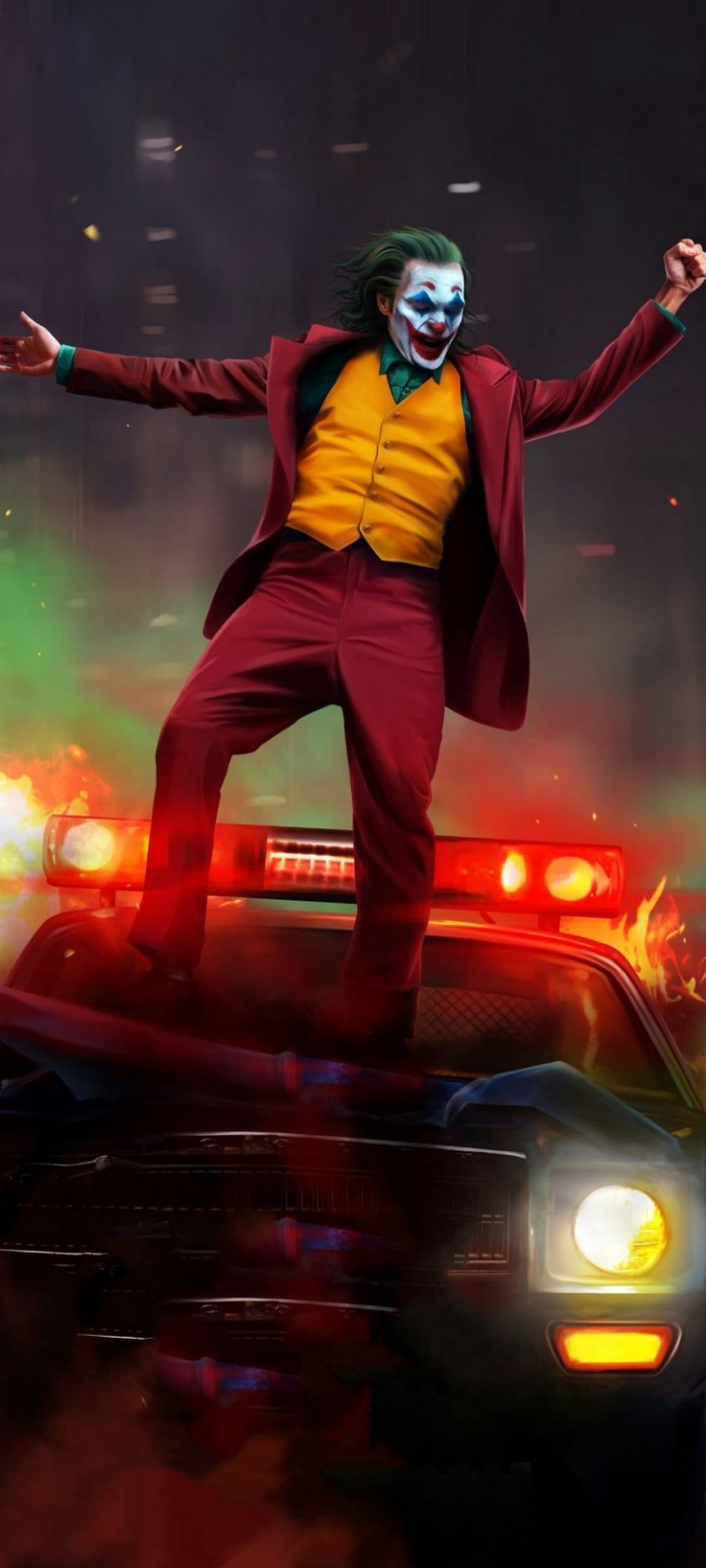 The Joker with green hair and clown makeup stands on a police car surrounded by orange flames and thick smoke.