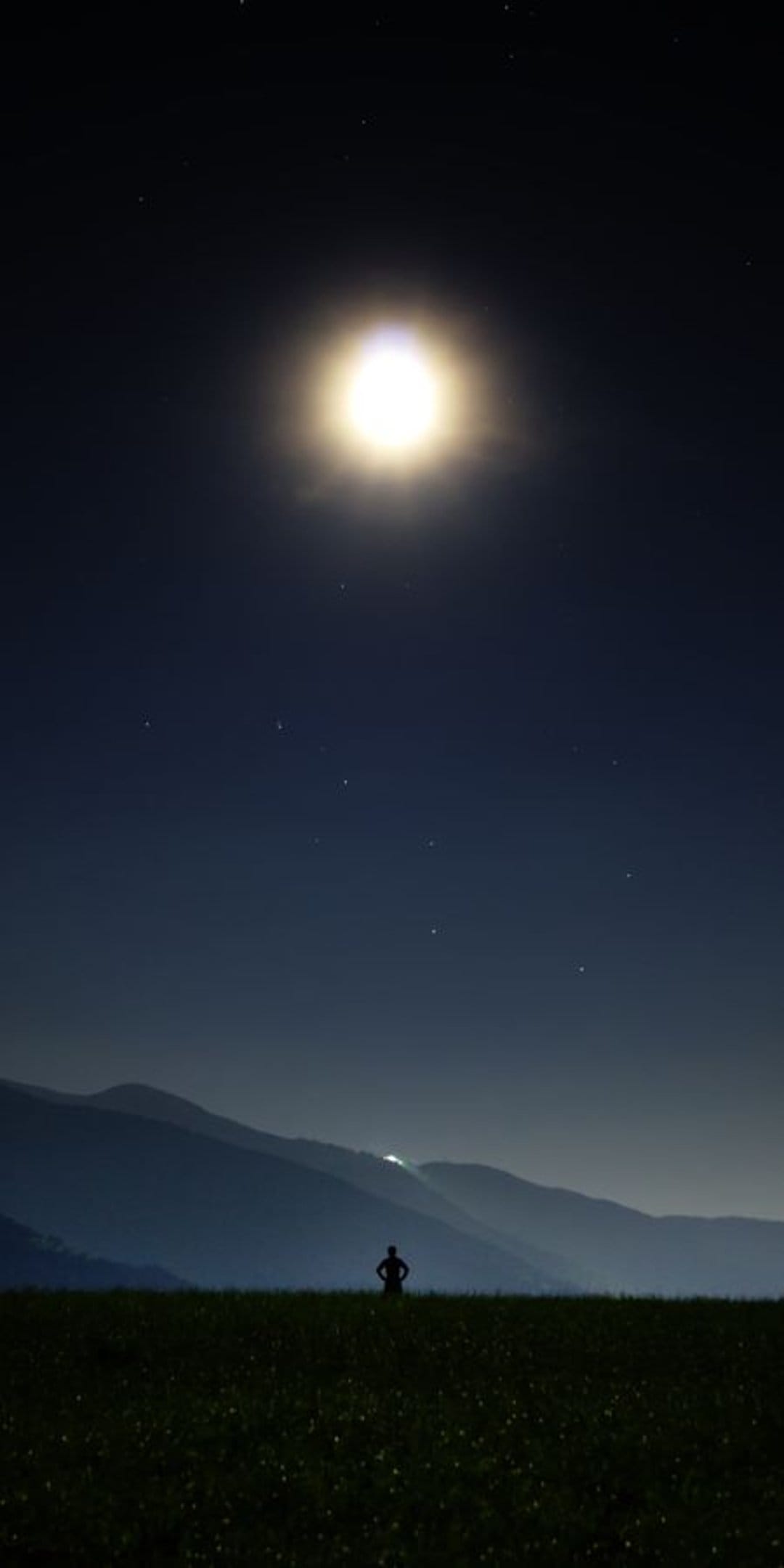 Lone person silhouette standing in a dark field beneath a massive glowing full moon and a starry night sky.