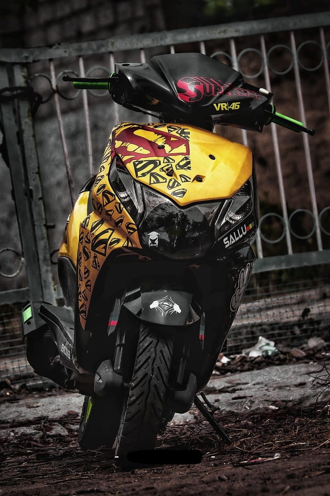 Yellow and black custom scooter with Superman emblems and VR46 decals parked against a metal fence outdoors.