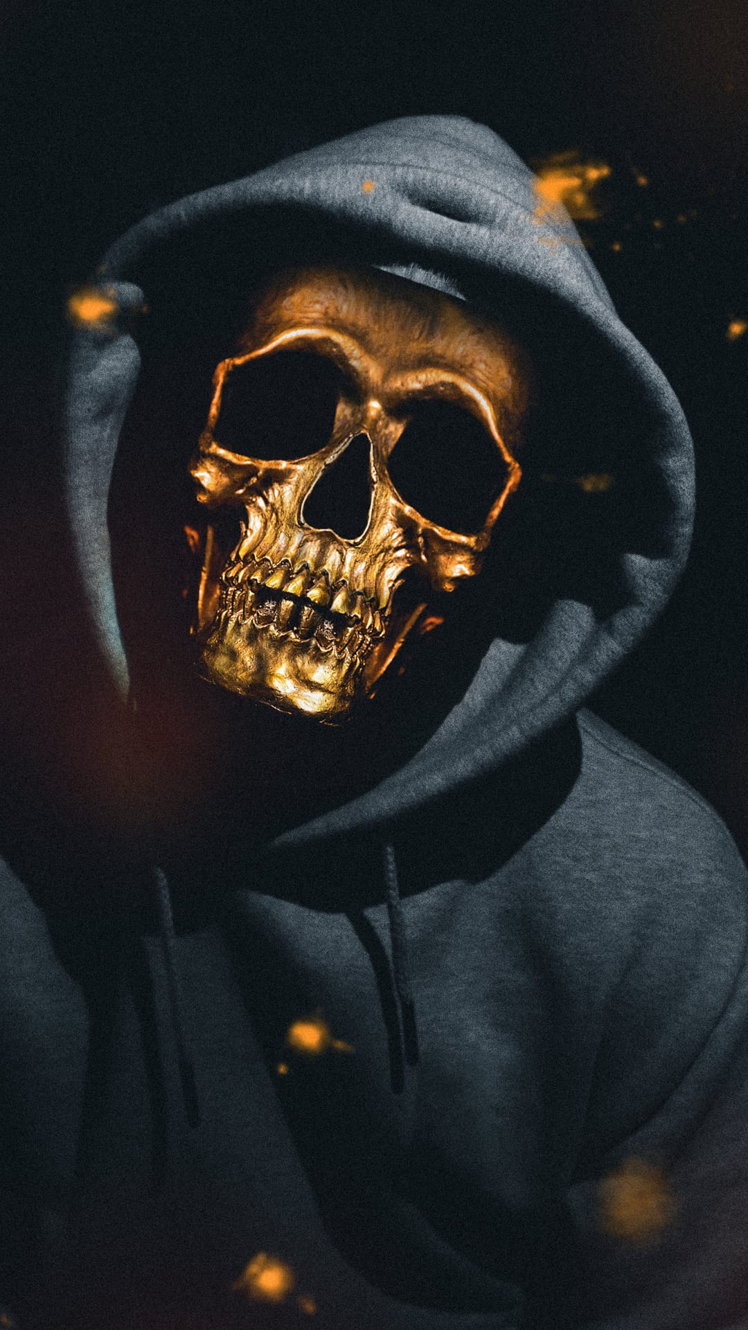 Golden skull mask worn under a dark hoodie surrounded by glowing orange embers and floating light particles.