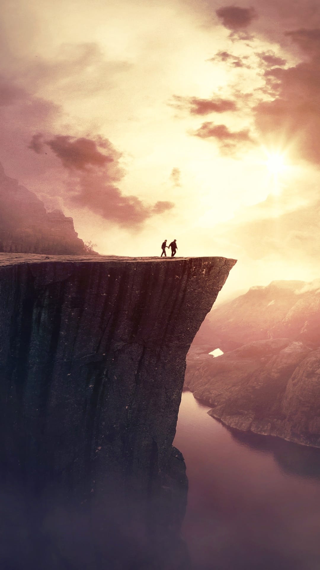 Two silhouetted figures hold hands on a steep cliff overlooking a canyon during a vibrant orange and purple sunset.