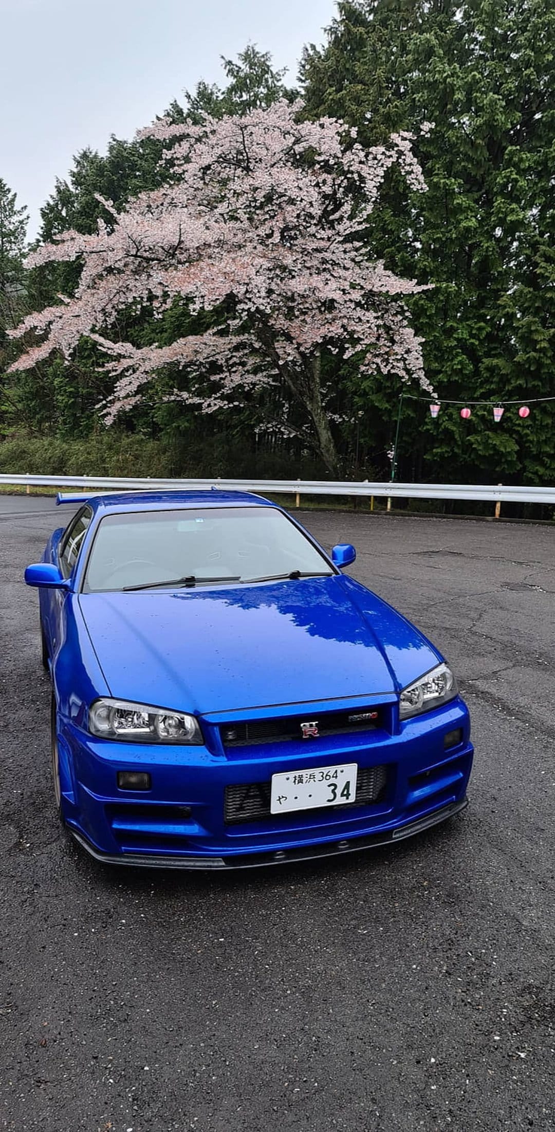 Nissan Skyline R34 GT-R in blue parked on a road under pink cherry blossoms with a large rear spoiler visible.