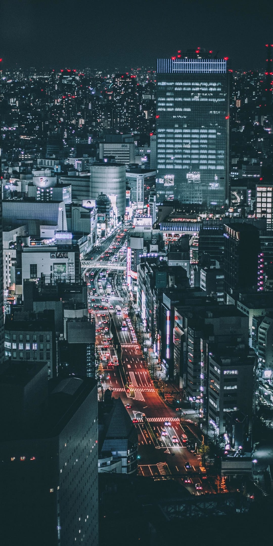 Tokyo skyscrapers glow with blue and magenta neon lights above busy transit arteries in a vertical night scene.