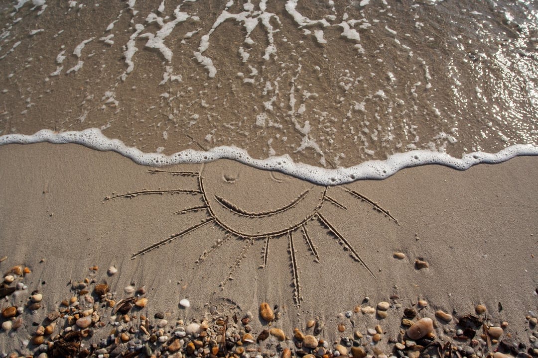 Smiling sun drawing in wet sand being washed over by white sea foam and gentle waves on a pebble-strewn beach.