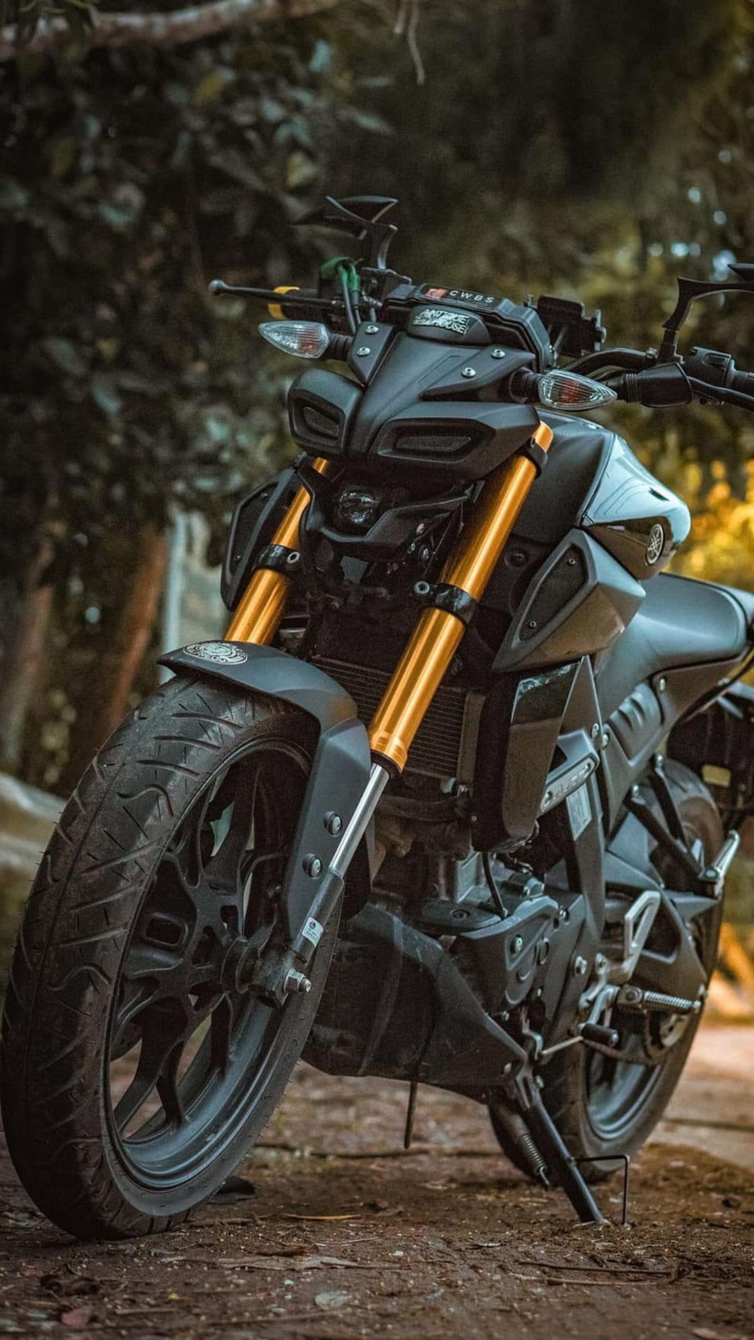 Yamaha MT-15 motorcycle featuring golden front forks and LED headlights parked against a green forest backdrop.