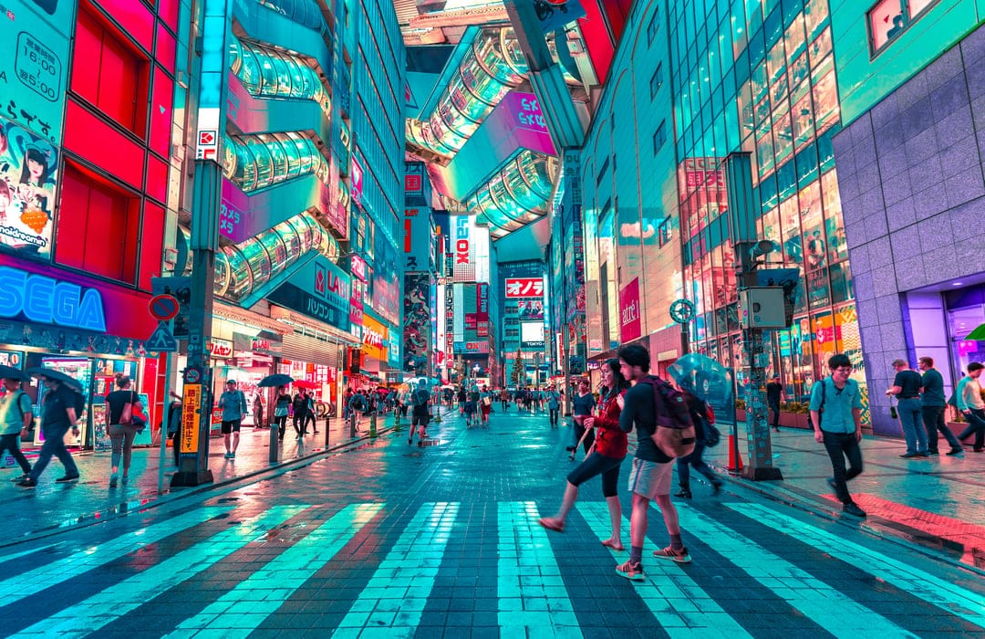 Pedestrians cross a white zebra-striped street illuminated by pink and blue neon building signs at night in Tokyo.