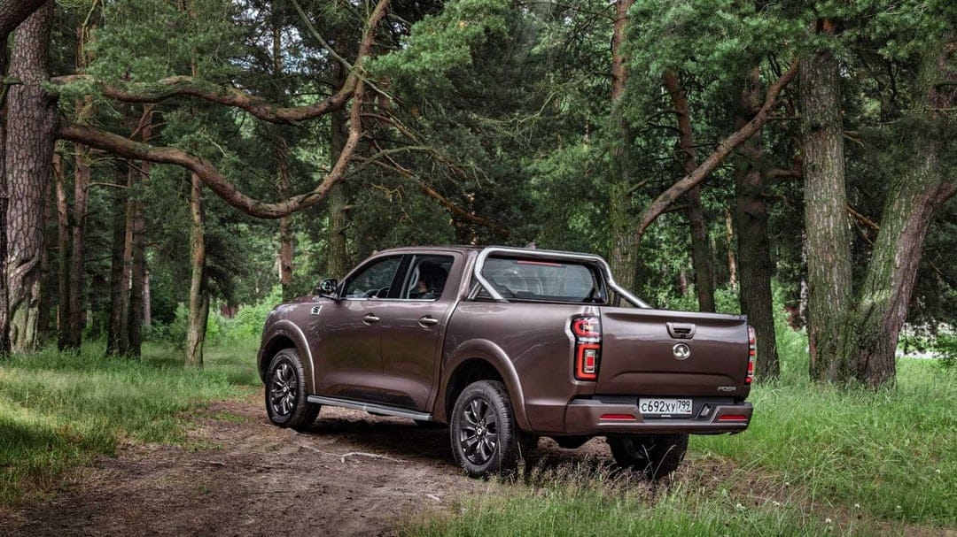 Brown pickup truck with chrome roll bar parked on a dirt path surrounded by tall green forest trees.