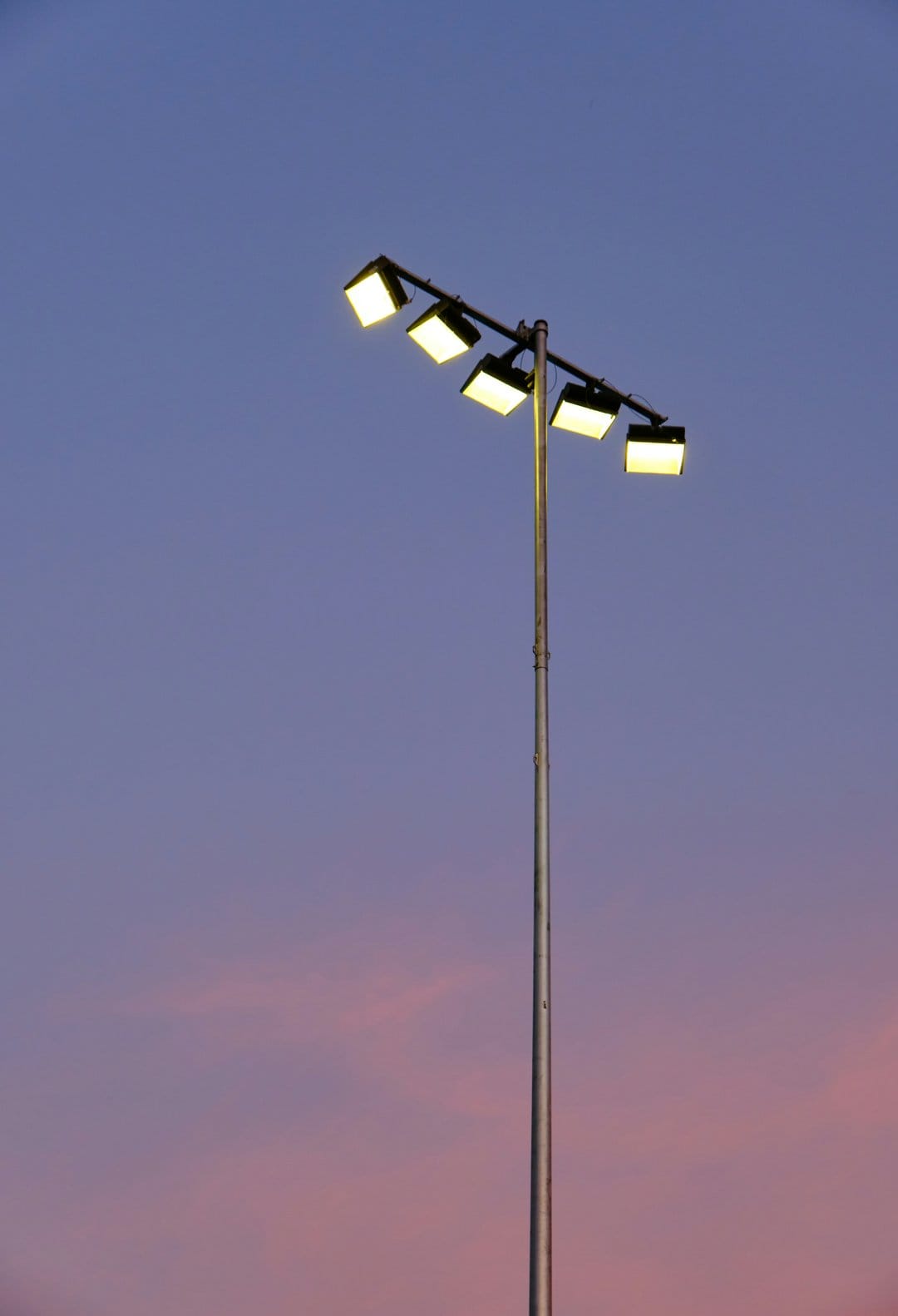 Metallic floodlight pole with glowing yellow lamps against a gradient sky of dark blue, purple, and soft pink.