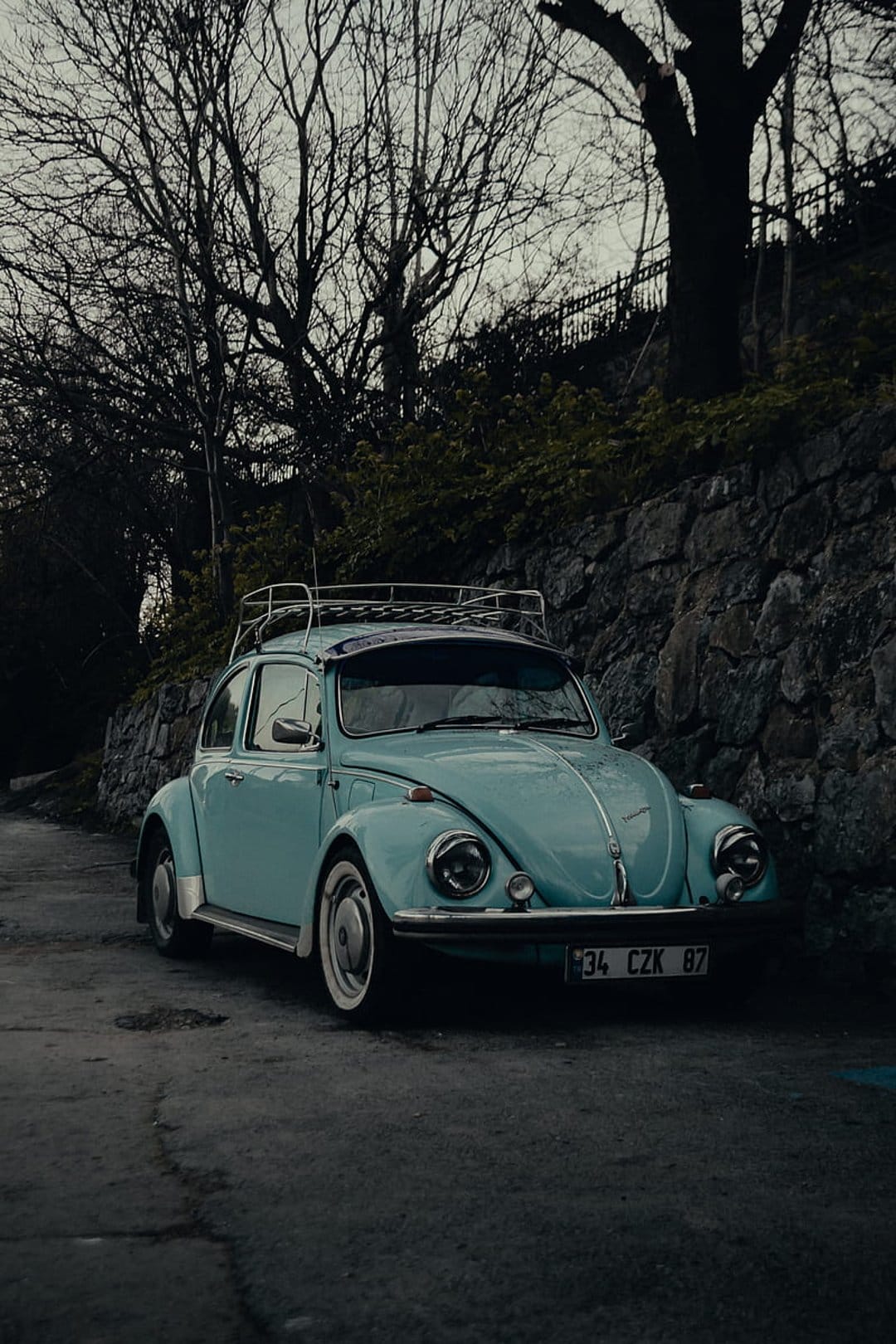 Turquoise vintage Volkswagen Beetle with a silver roof rack parked beside a stone wall against dark tree silhouettes.