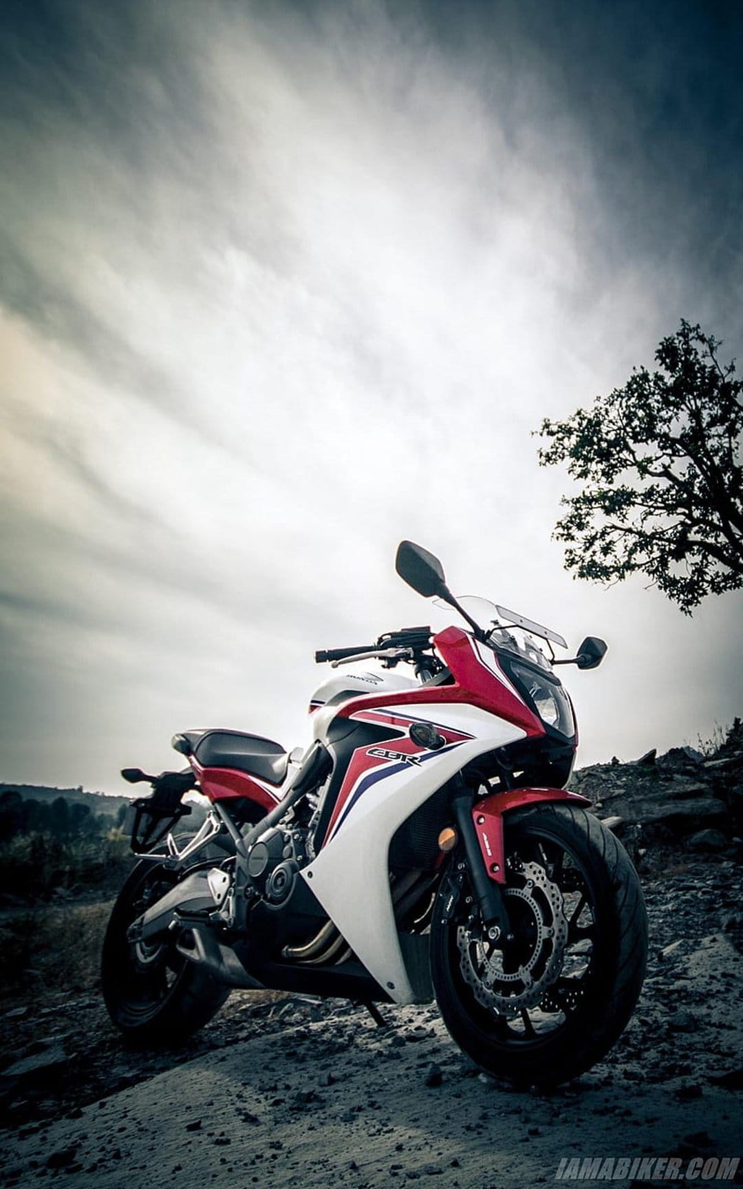 Red and white Honda CBR sportbike parked on a rocky cliffside under dark clouds with a silhouetted tree.