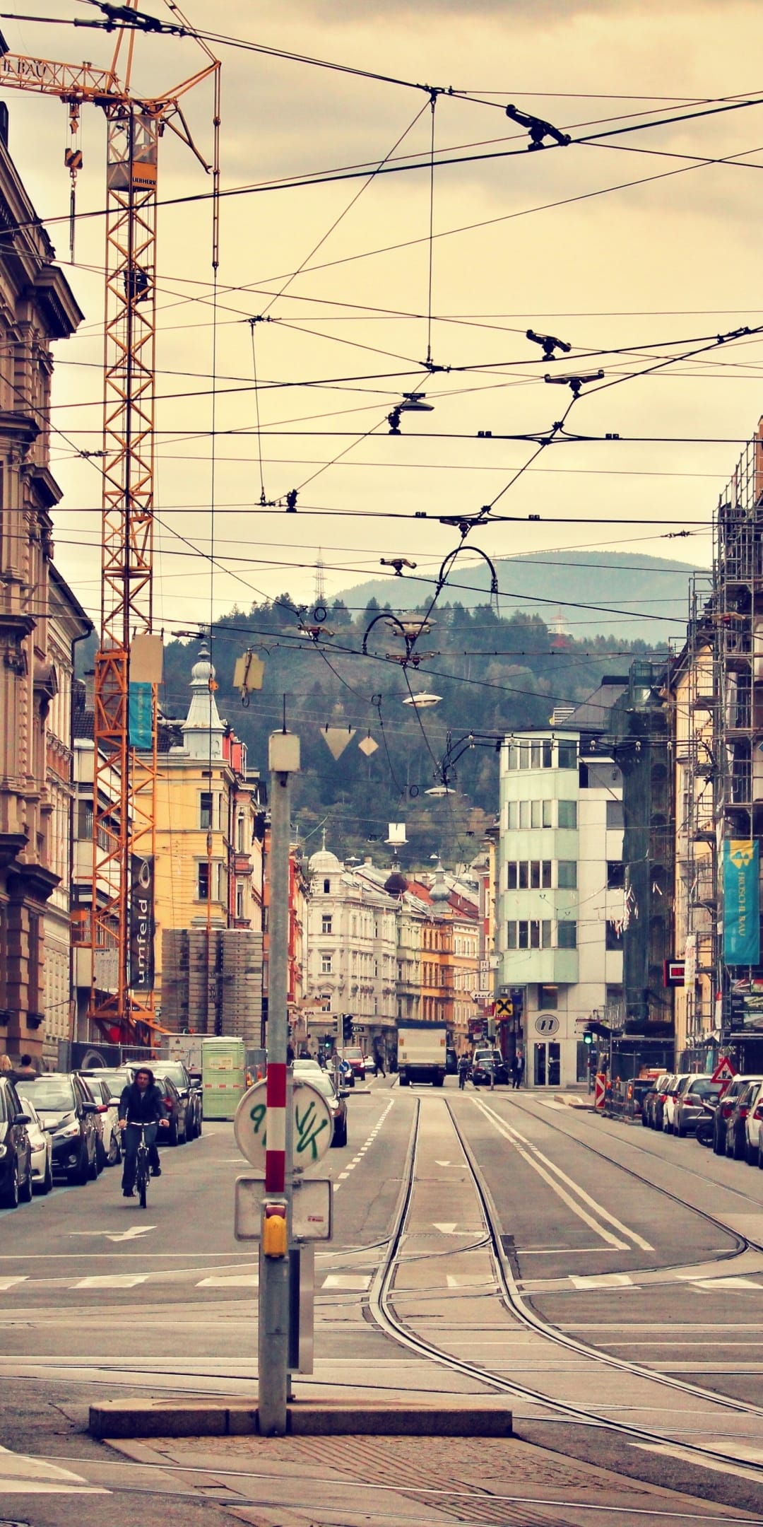 A yellow construction crane stands beside classic buildings and a cyclist on an Innsbruck street with mountains.