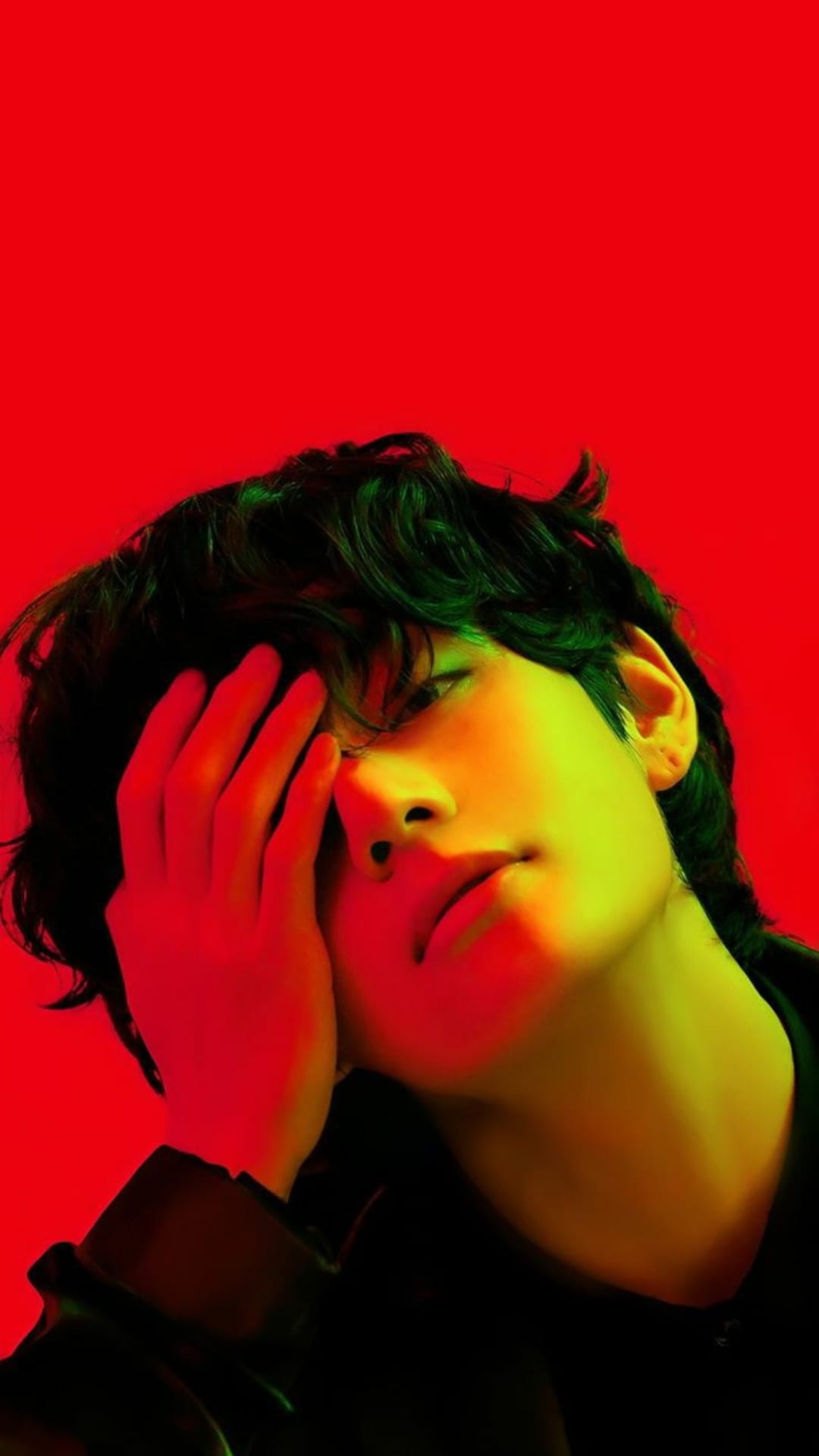 Young man with wavy hair obscuring his face with a hand under intense red and yellow gradient studio lighting.