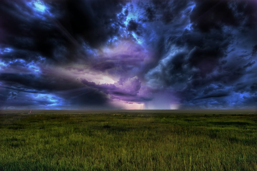 Swirling purple and blue storm clouds massing above a wide green meadow under a dark, heavy sky during a tempest.