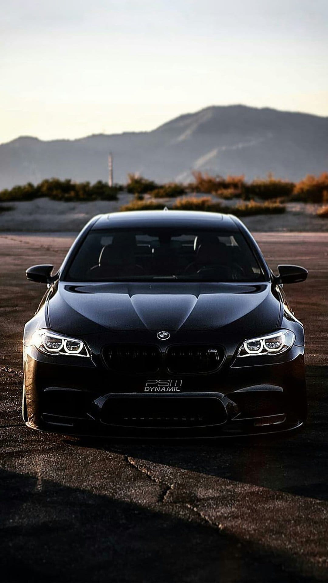 Black BMW M5 with glowing headlights parked on a sandy desert road under a hazy, dusky sky during twilight.