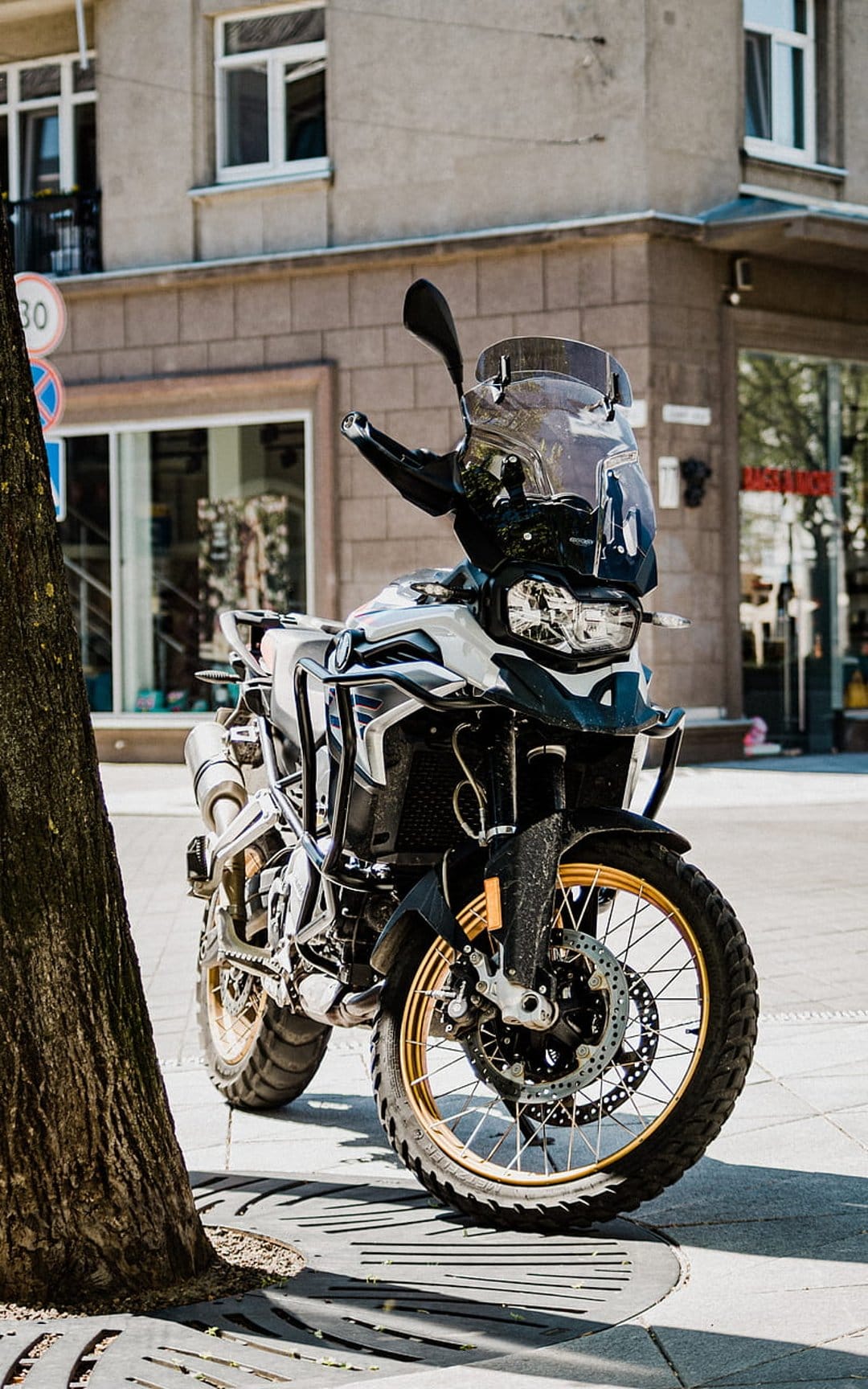 BMW adventure motorcycle with gold rims parked on a city sidewalk next to a tree trunk and urban storefront.