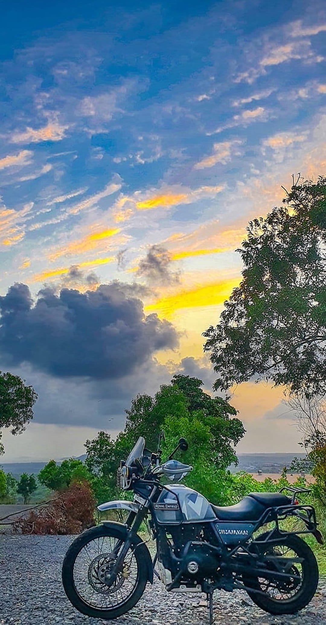Royal Enfield Himalayan motorcycle on a gravel road with a vibrant orange and blue sunset sky and green trees.