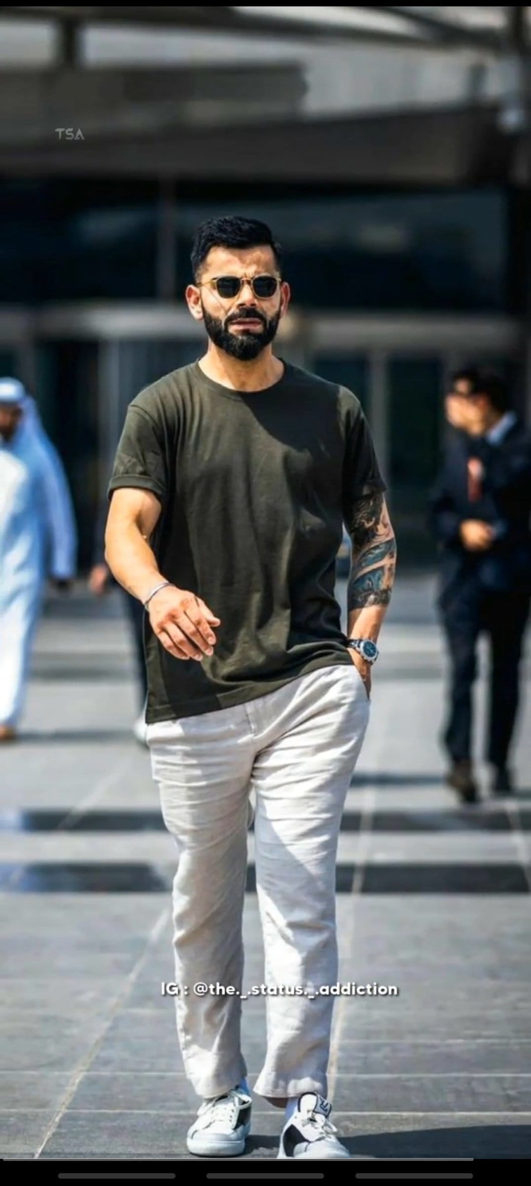 Virat Kohli walking in a green shirt and sunglasses with visible arm tattoos and a blurred street background.