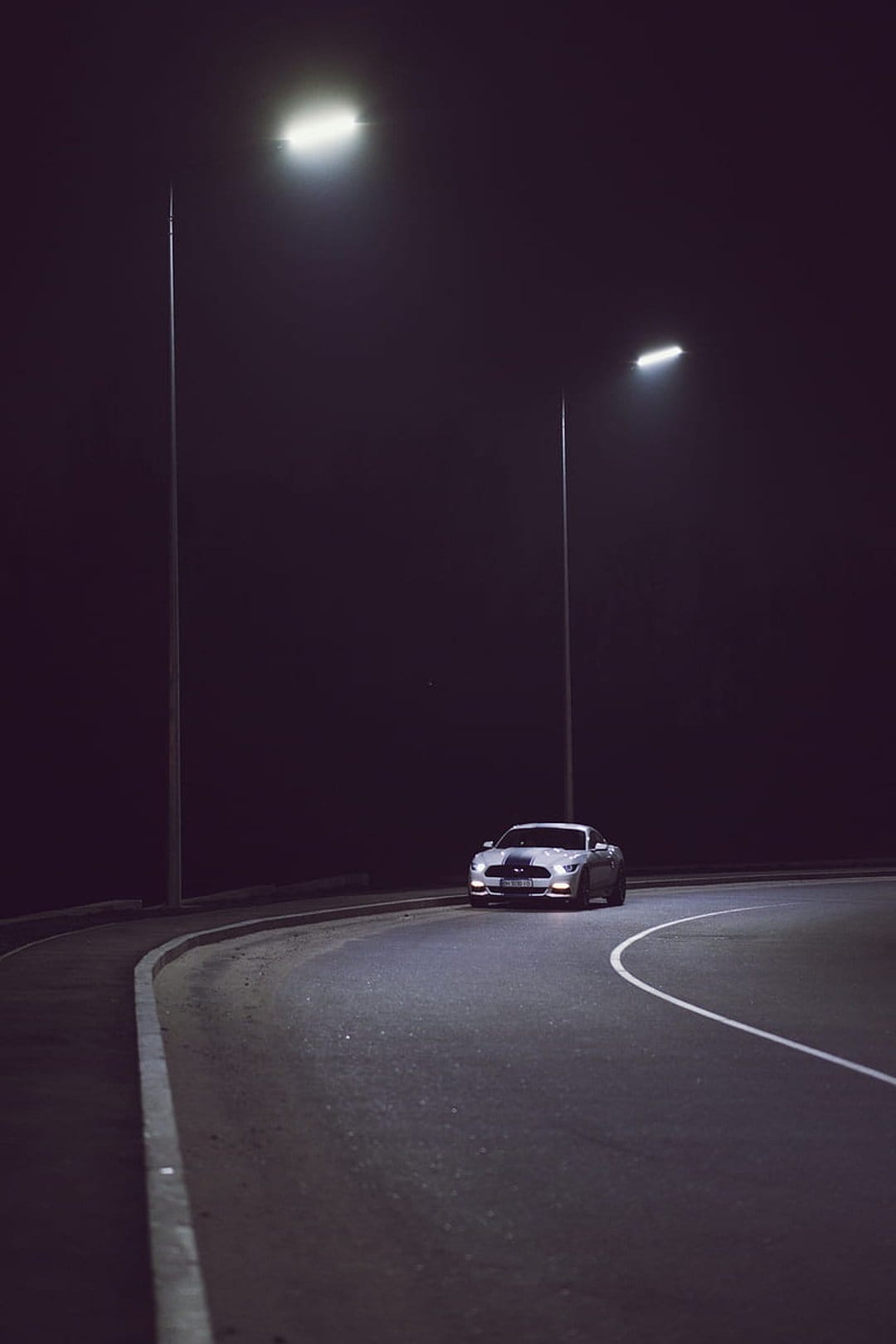 White Ford Mustang sports car cruising a dark curved road at night illuminated by overhead street lamps.