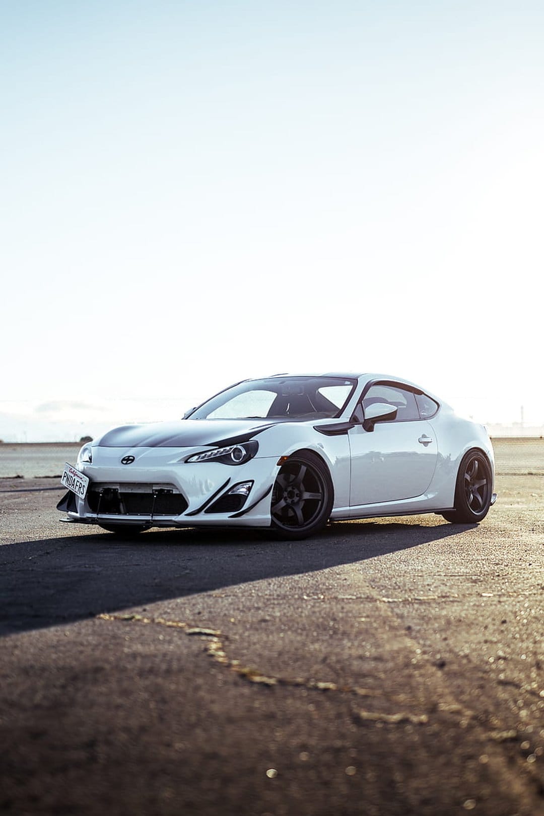 Toyota GT86 with a white paint job and black hood accents parked on dark asphalt under a bright clear sky.