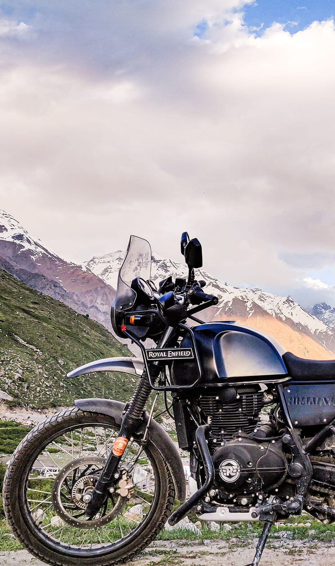 Royal Enfield motorcycle parked on a green hill facing snow-capped mountains under a cloudy, overcast sky.