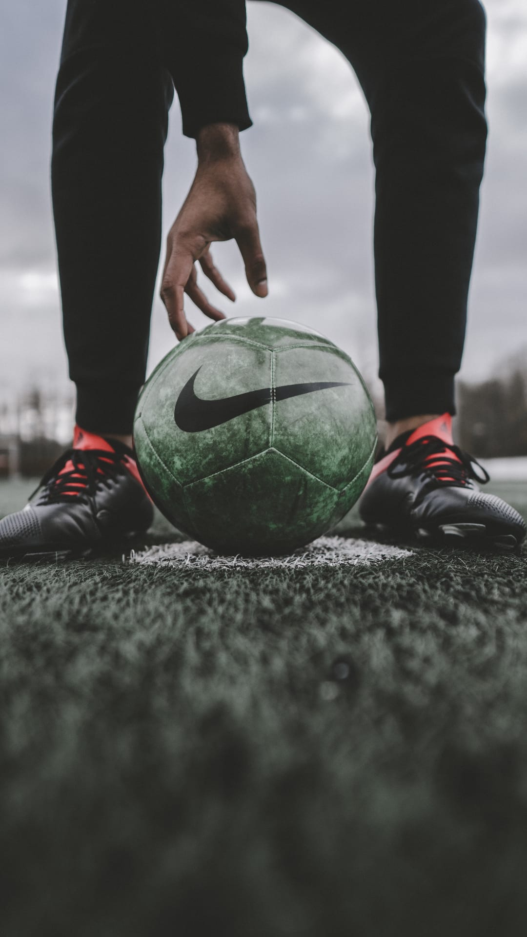 Soccer player's hand reaching toward a green Nike ball on a grassy pitch with red-accented cleats visible.