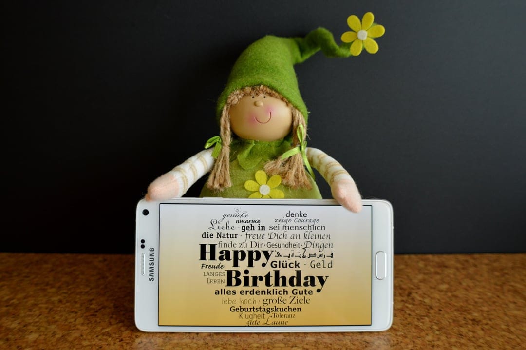 Felt elf in a green pointed hat holds a smartphone with birthday typography on a dark wood background texture.