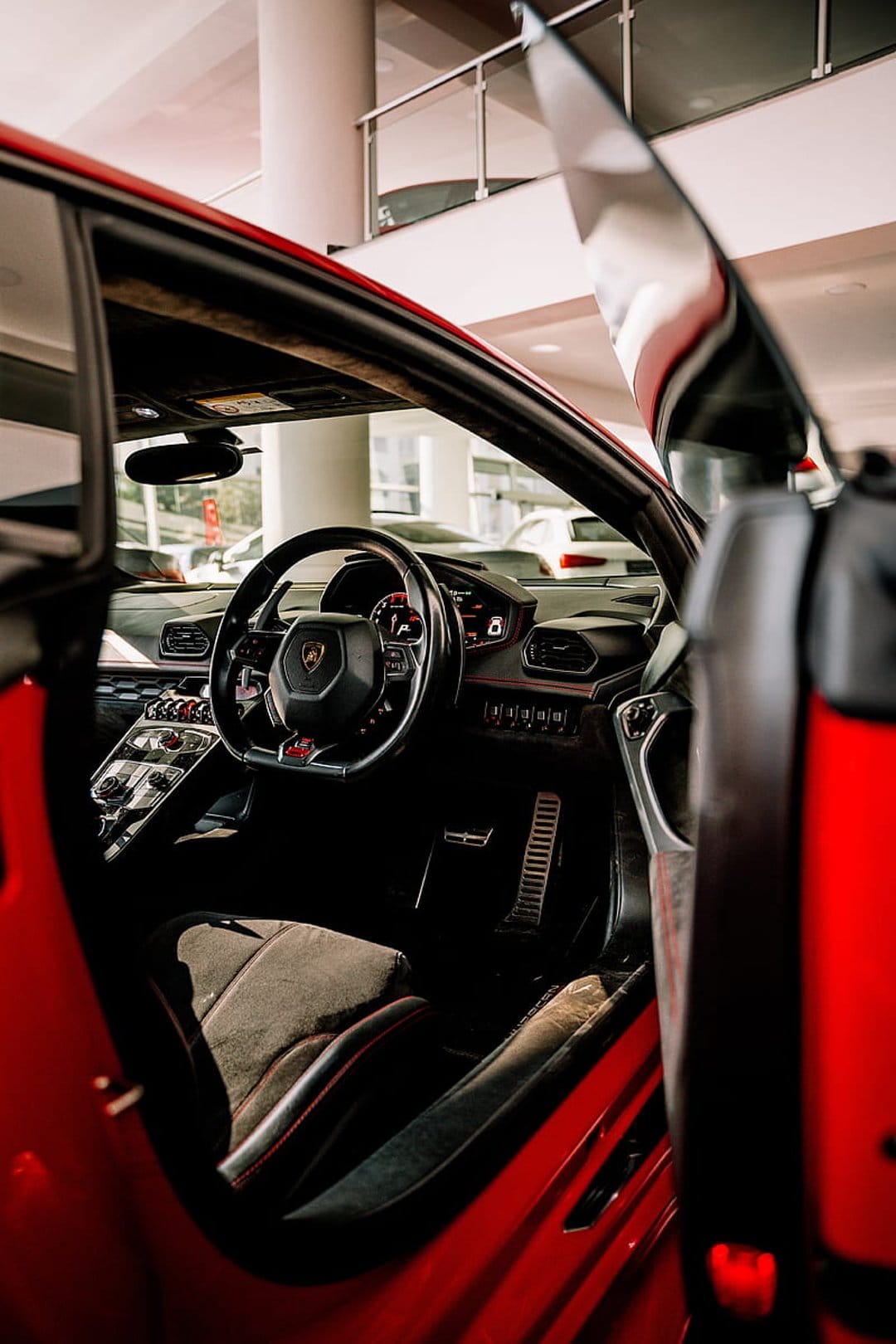 Red Lamborghini driver's seat and steering wheel framed by an open door in a vertical luxury car perspective.