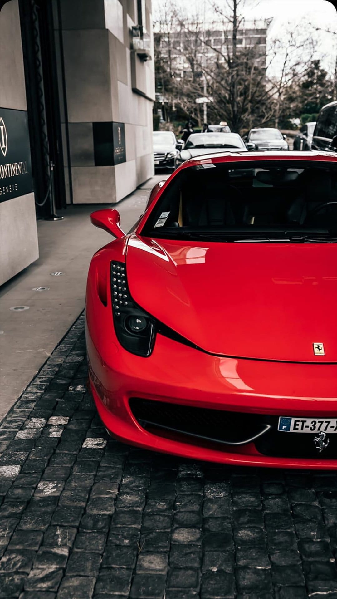Red Ferrari supercar with glowing headlights parked on a grey cobblestone street in front of a modern hotel.