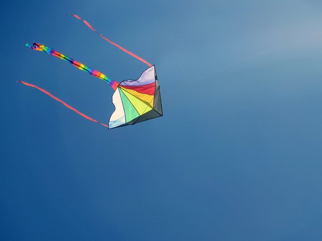 Rainbow diamond kite with red streamers flies in a clear blue sky under a soft, glowing midday sun.