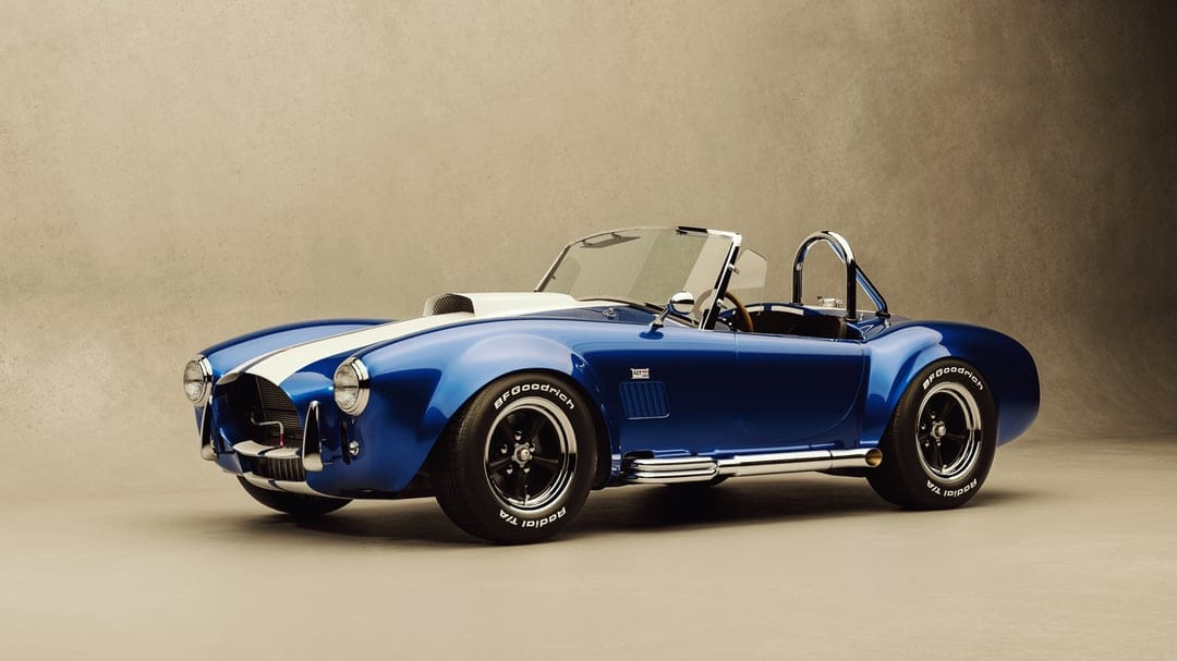 Blue Shelby Cobra sports car with white racing stripes and chrome side pipes against a beige studio background.