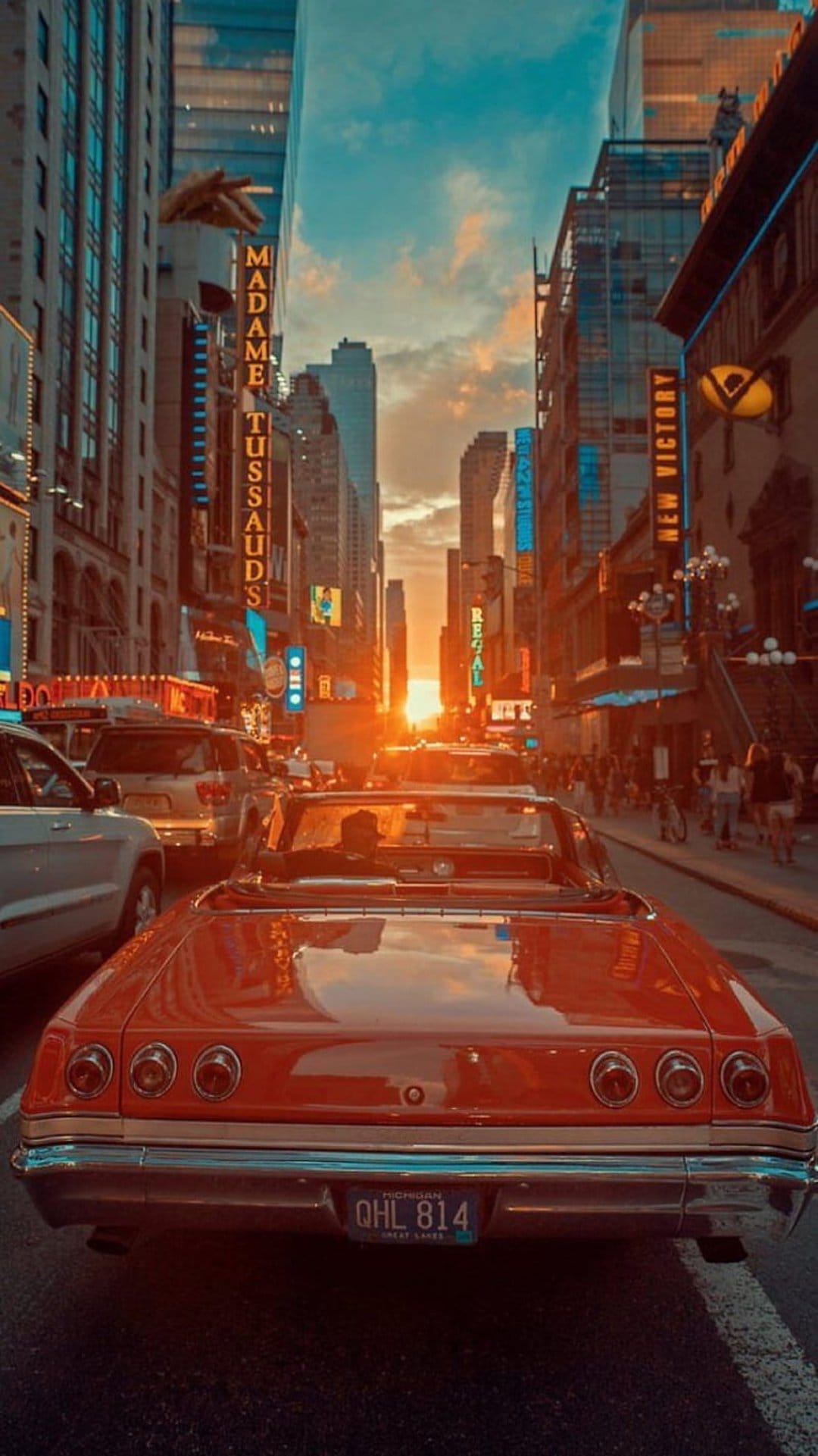 Red vintage convertible drives through Times Square at sunset with bright neon theater signs and golden light.