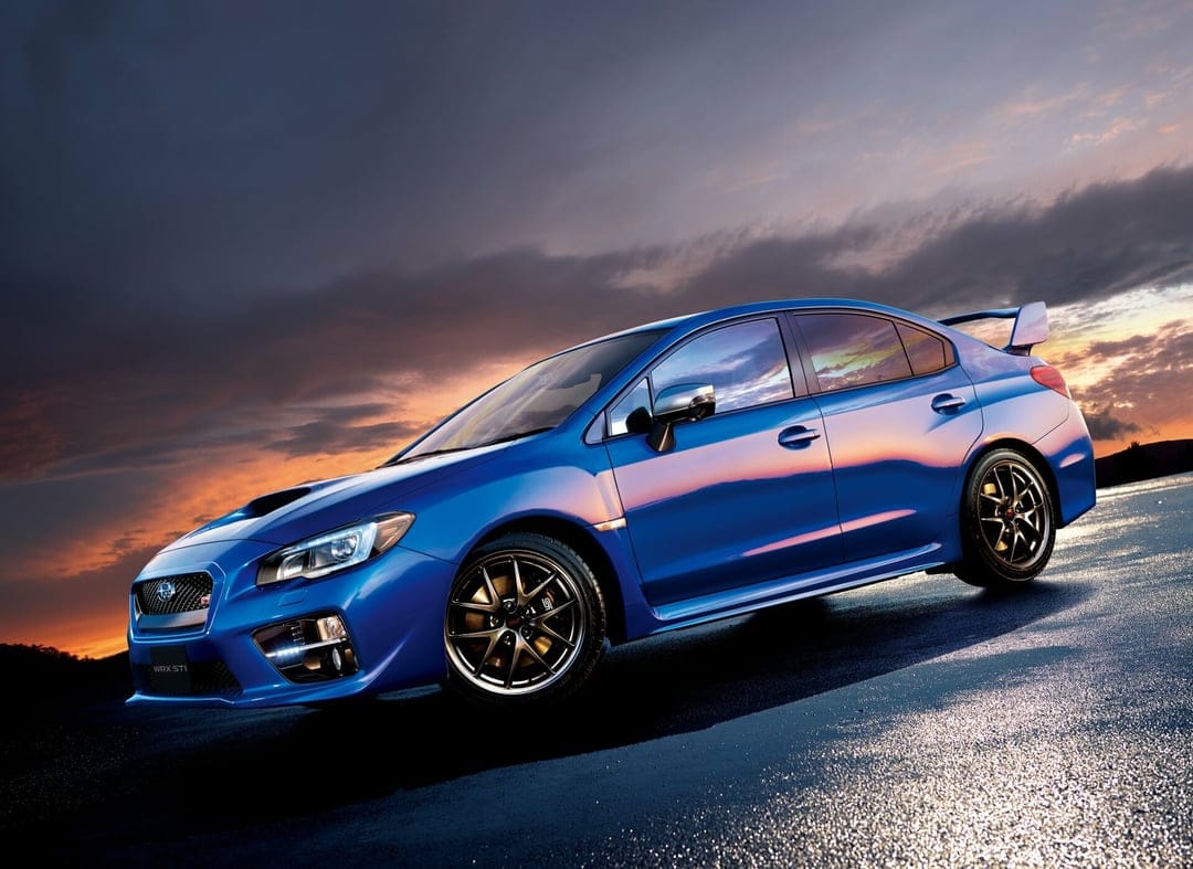 Blue Subaru WRX STI sports car with bronze wheels on wet asphalt under a vibrant orange and purple sunset sky.