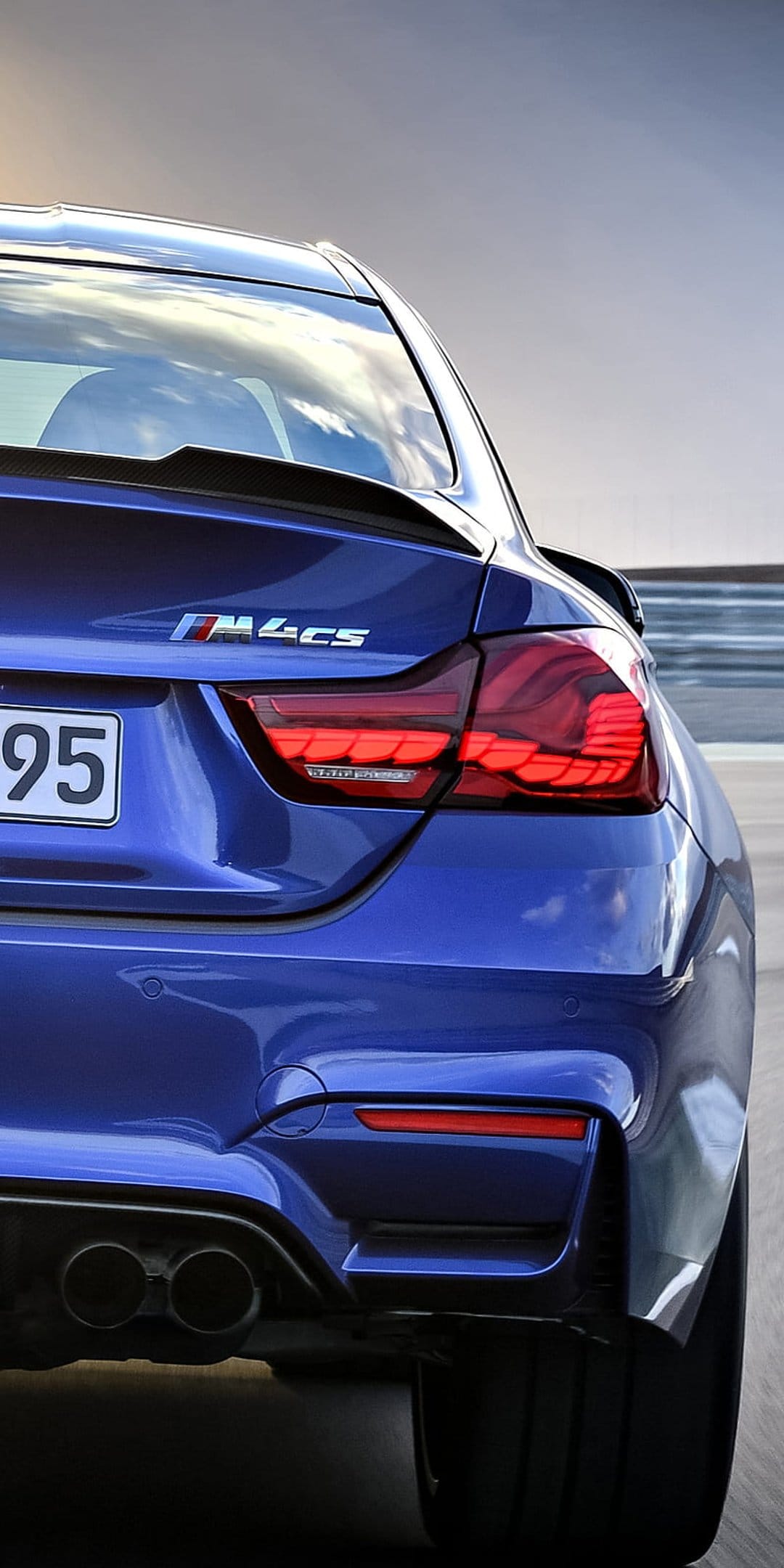 BMW M4 CS rear view featuring glowing LED taillights and quad exhaust pipes against a metallic blue paint finish.