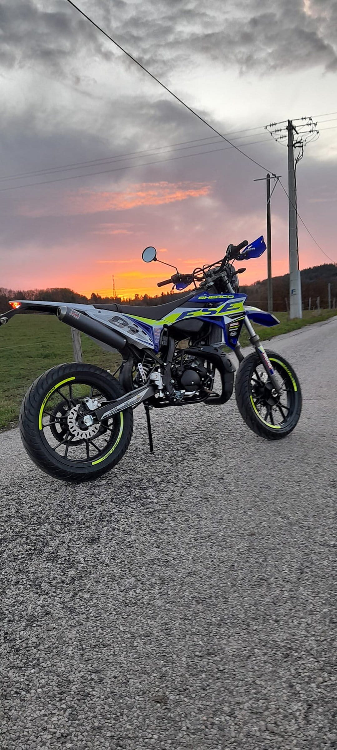 Blue and yellow dirt bike parked on a gravel road against a dramatic orange and pink sunset with power lines.