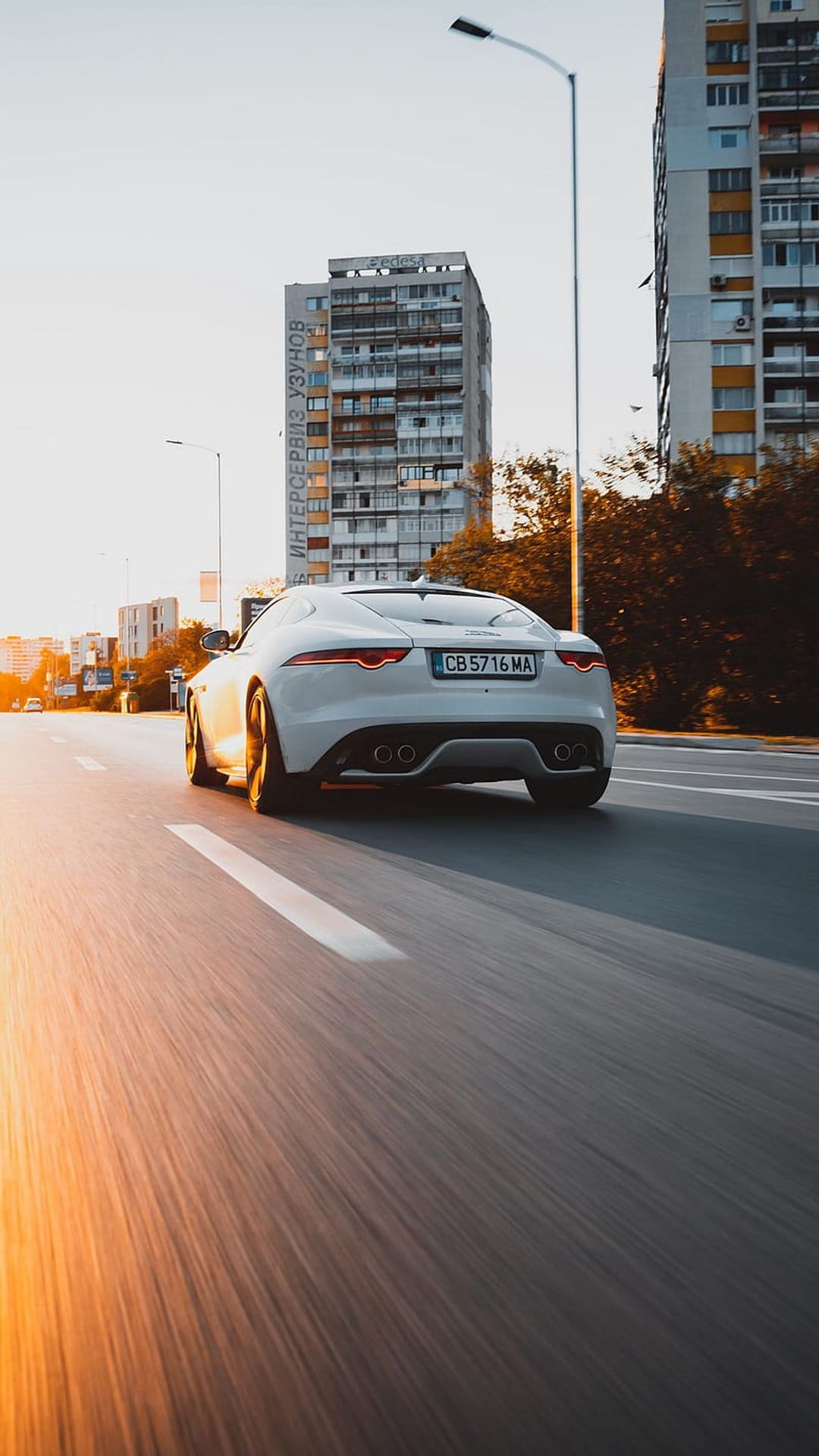 White Jaguar F-Type convertible driving on an urban road at sunset with motion blur and golden hour lighting.
