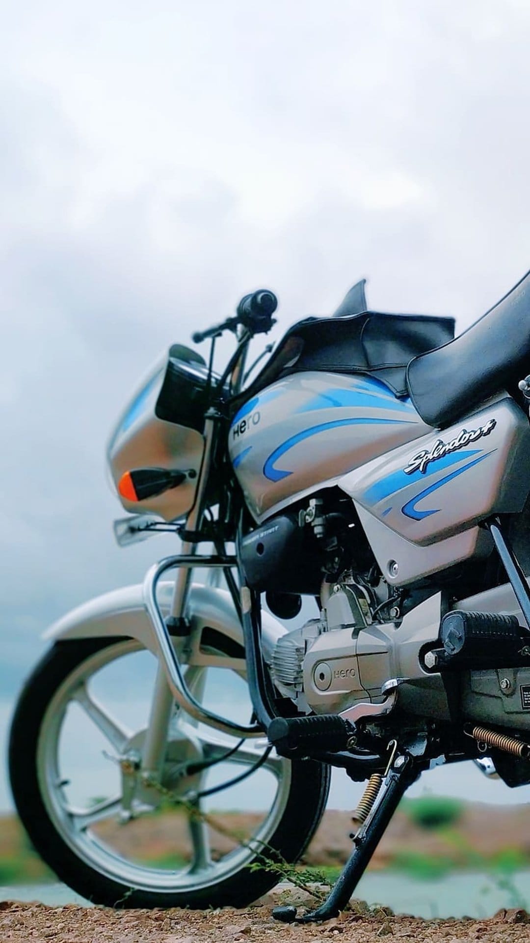 Silver Hero Splendor motorcycle engine and front wheel in a low-angle shot against a blurred green background.