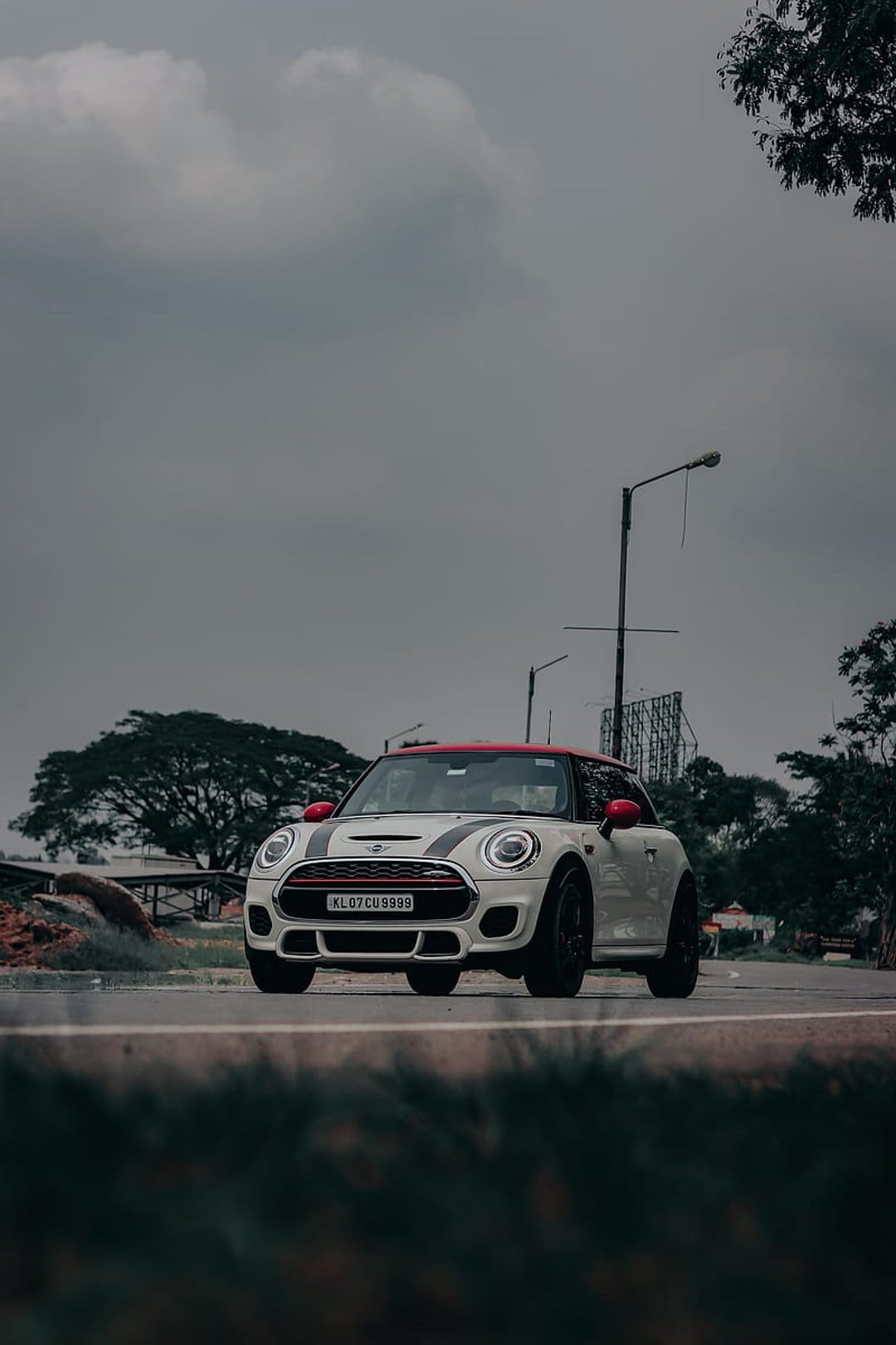 White Mini Cooper S with a red roof and racing stripes travels on a wet road under heavy, dark storm clouds.