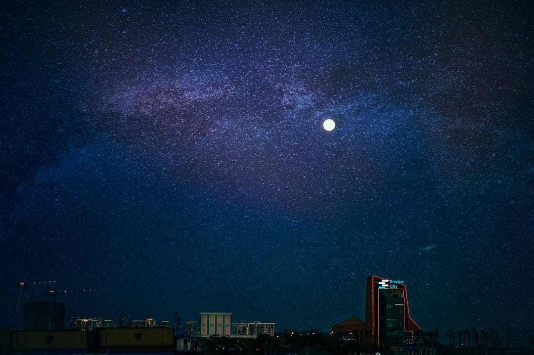 Illuminated skyscrapers stand beneath the swirling Milky Way galaxy and a bright moon in a deep purple night sky.