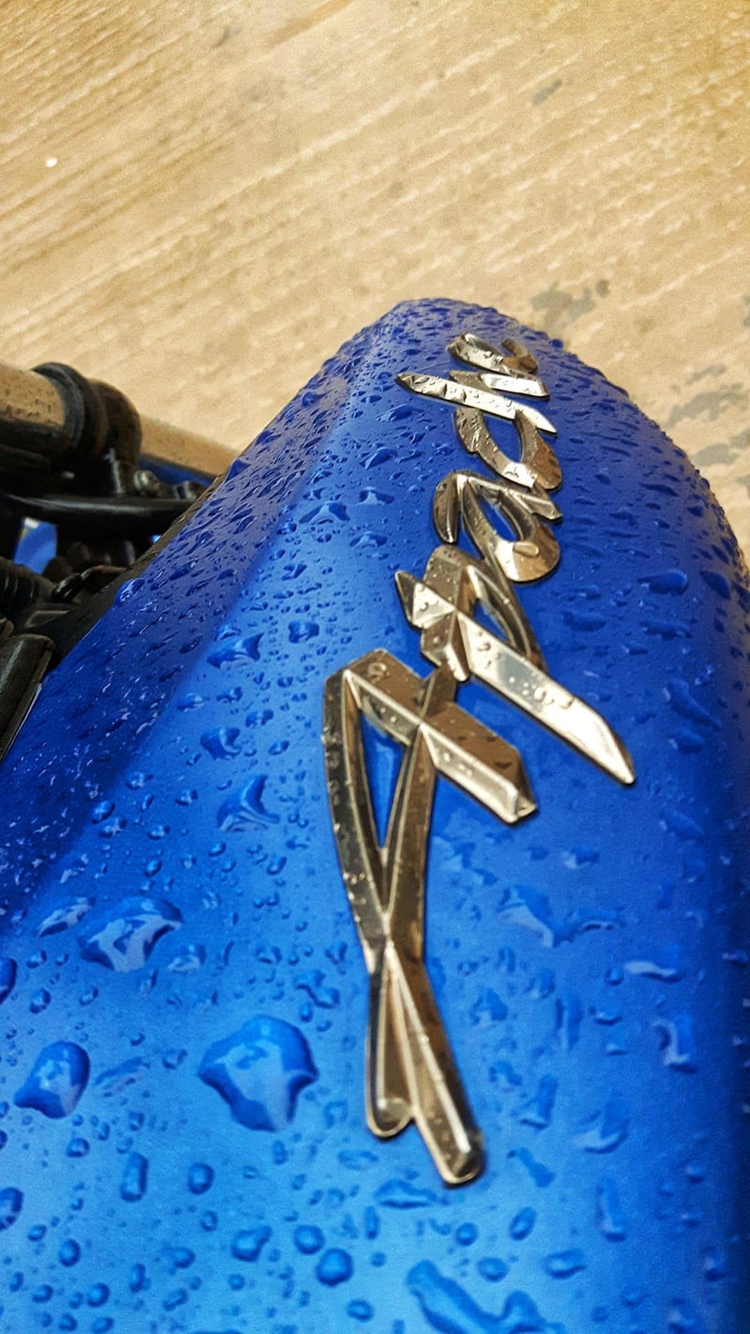 Blue motorcycle fuel tank with a metallic RX emblem and scattered water droplets against a wooden background.