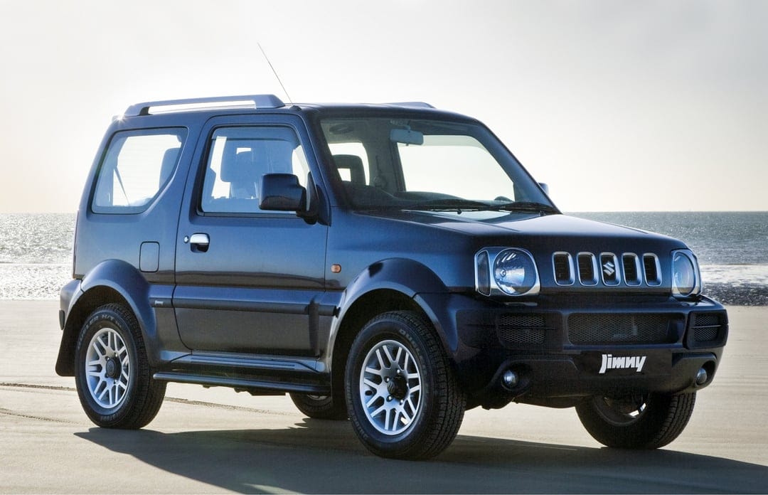 Dark blue Suzuki Jimny SUV parked on a sandy beach with a soft ocean horizon and bright sky in the background.