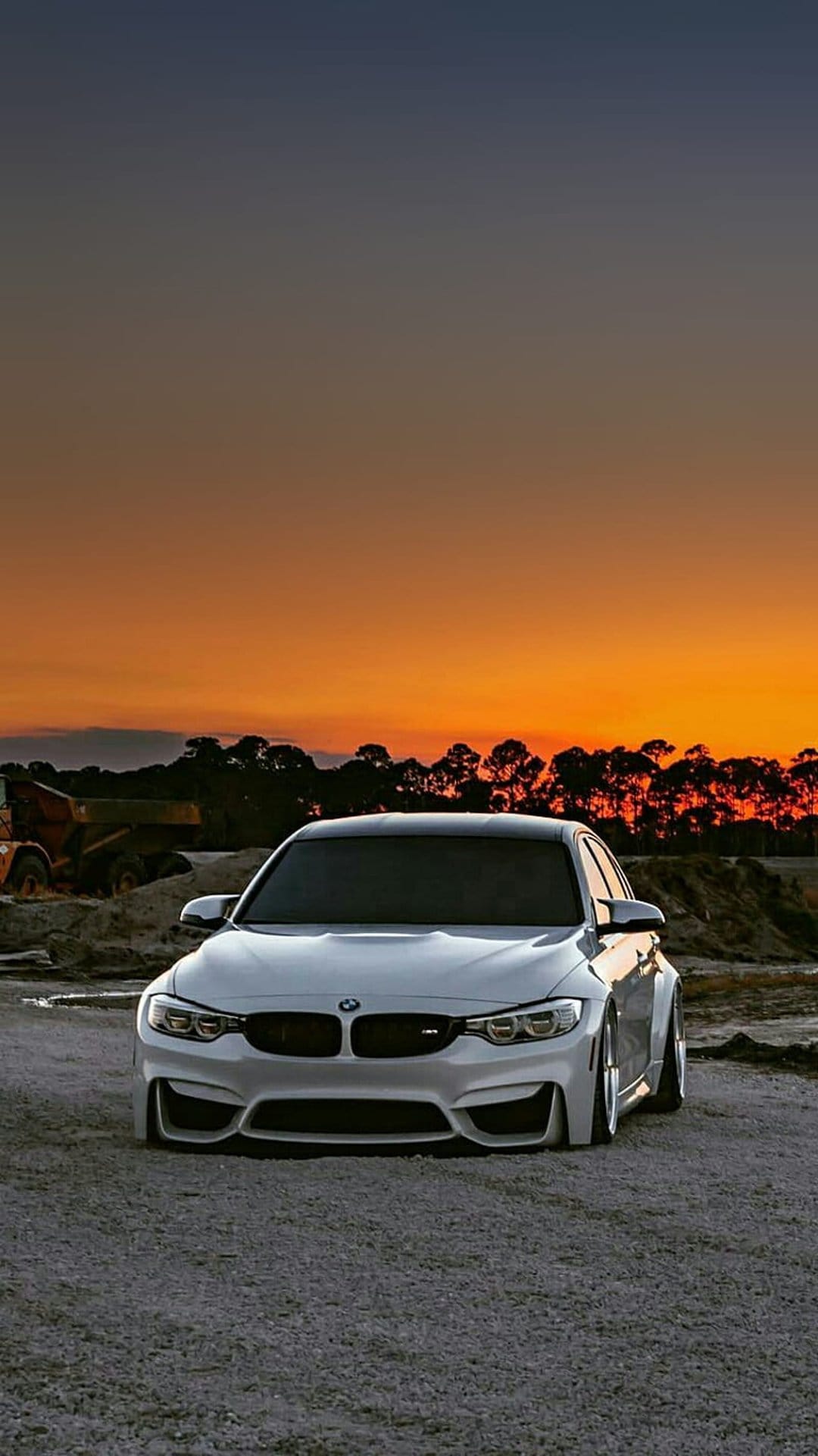 White BMW M4 parked on gravel with heavy construction machinery under a glowing orange and yellow sunset sky.