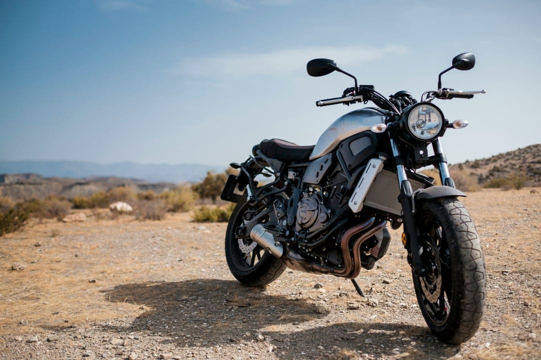 Silver Yamaha motorcycle parked on sandy desert terrain with rolling hills under a vibrant, clear blue sky.