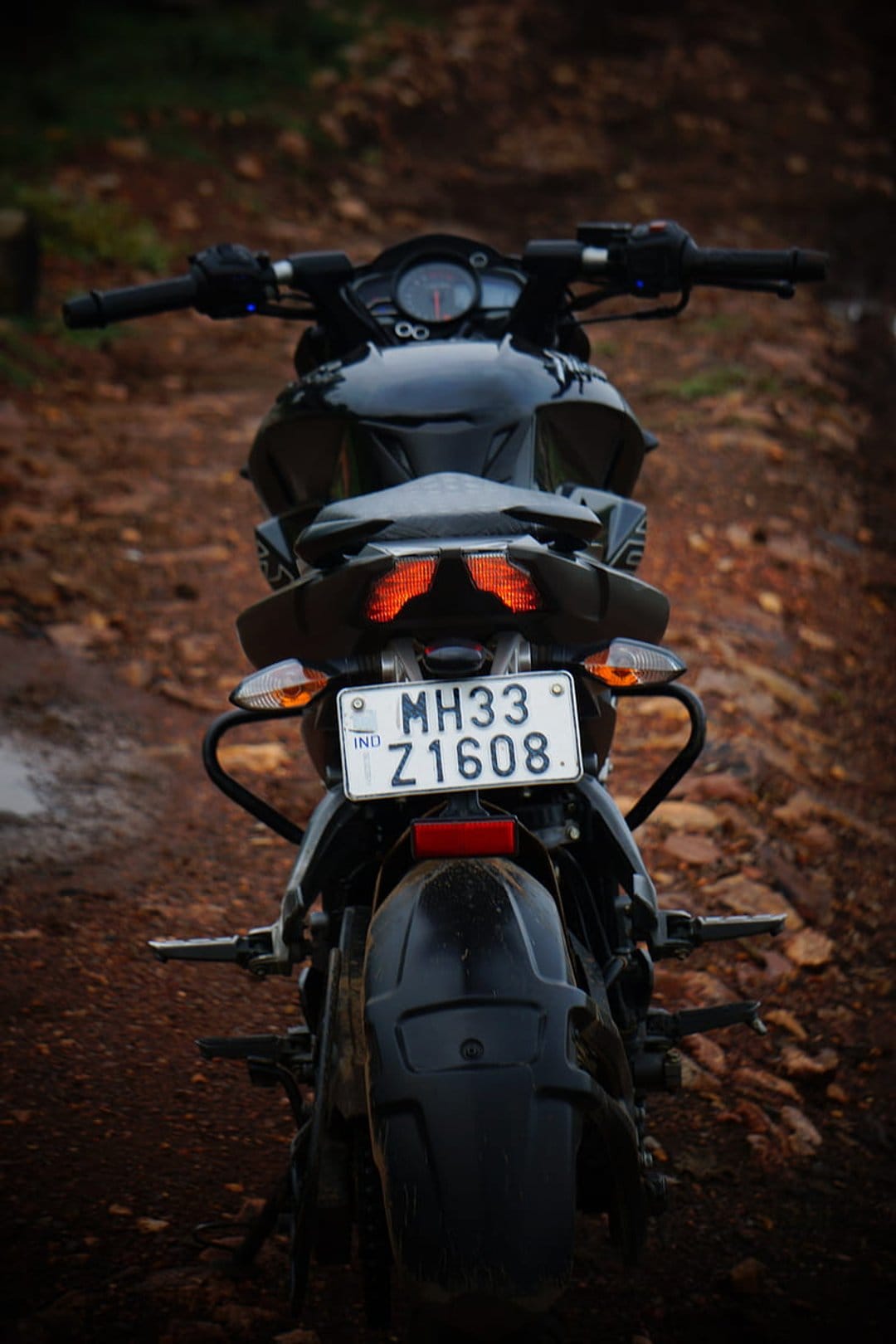 Black motorcycle rear fender with orange lights and a license plate on a gravel road with a blurred backdrop.