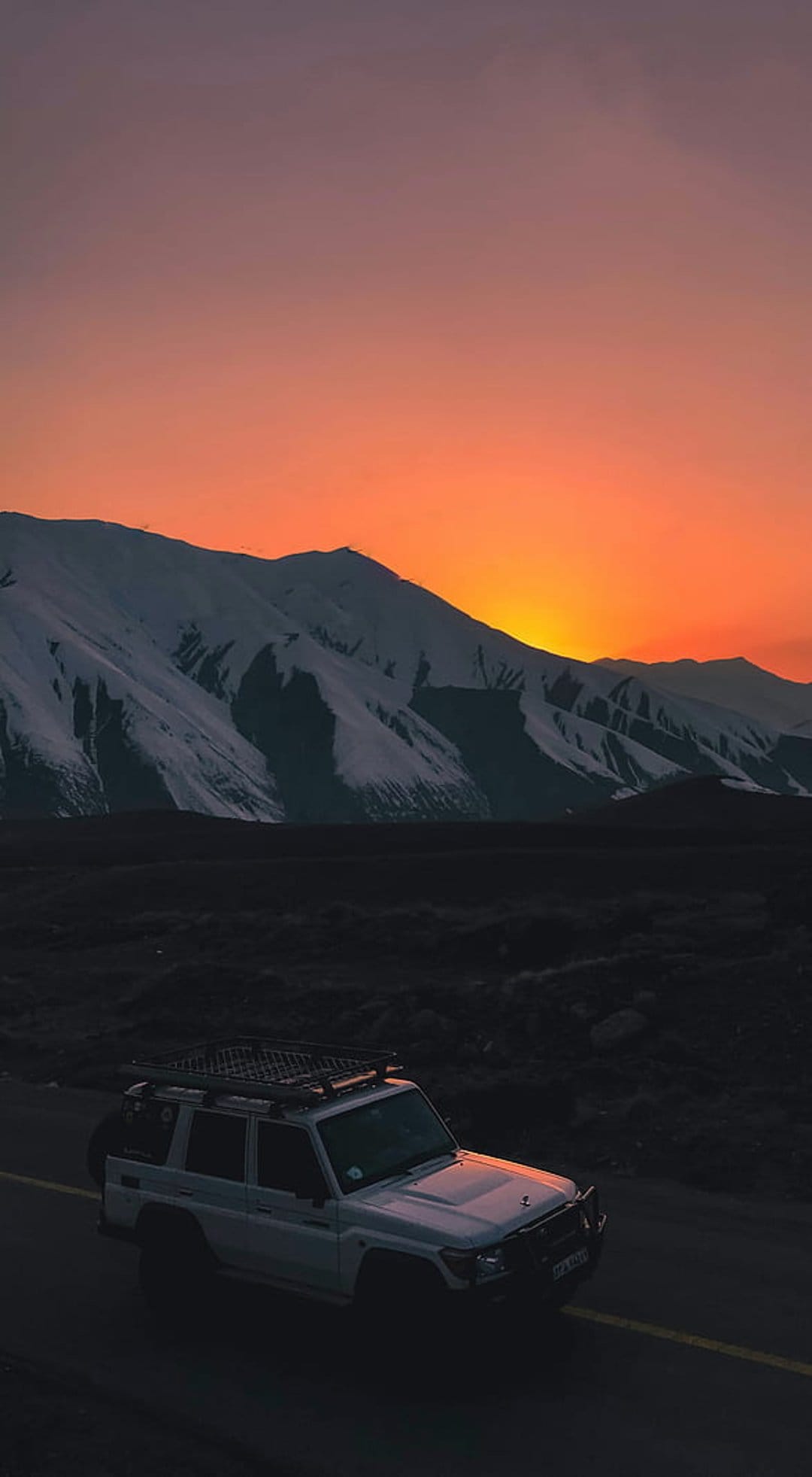 White SUV with roof rack on a mountain road before snow peaks under an orange and violet sunset sky.