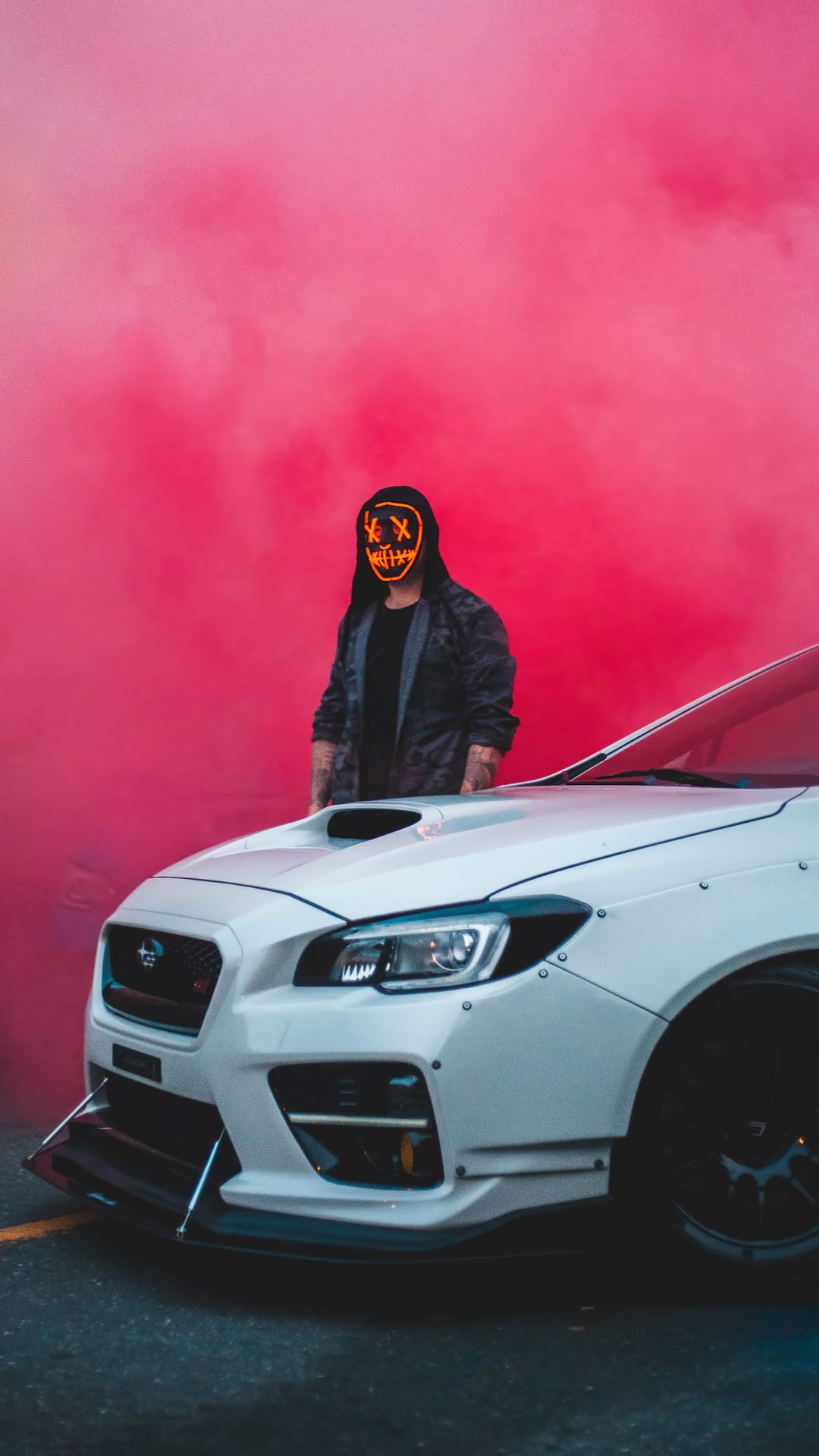 Masked person in a black hoodie stands near a white sports car surrounded by thick clouds of red smoke at night.