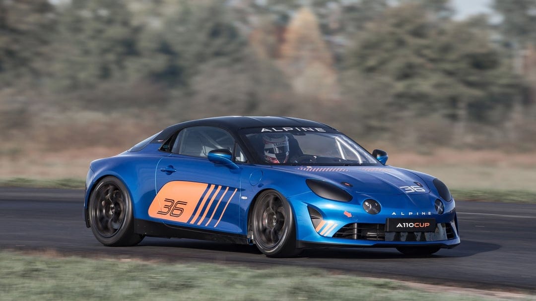 Blue Alpine A110 Cup race car with orange decals speeding on an asphalt track with motion blur background.