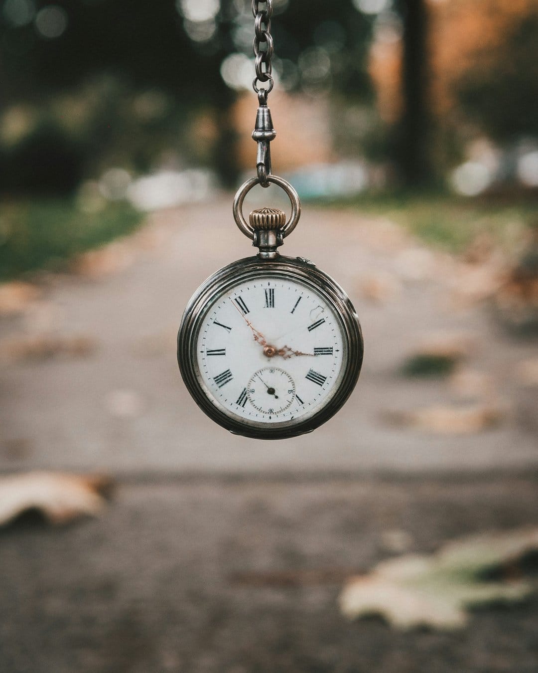 Silver pocket watch with Roman numerals hanging from a chain over a blurred autumn path with golden fallen leaves.