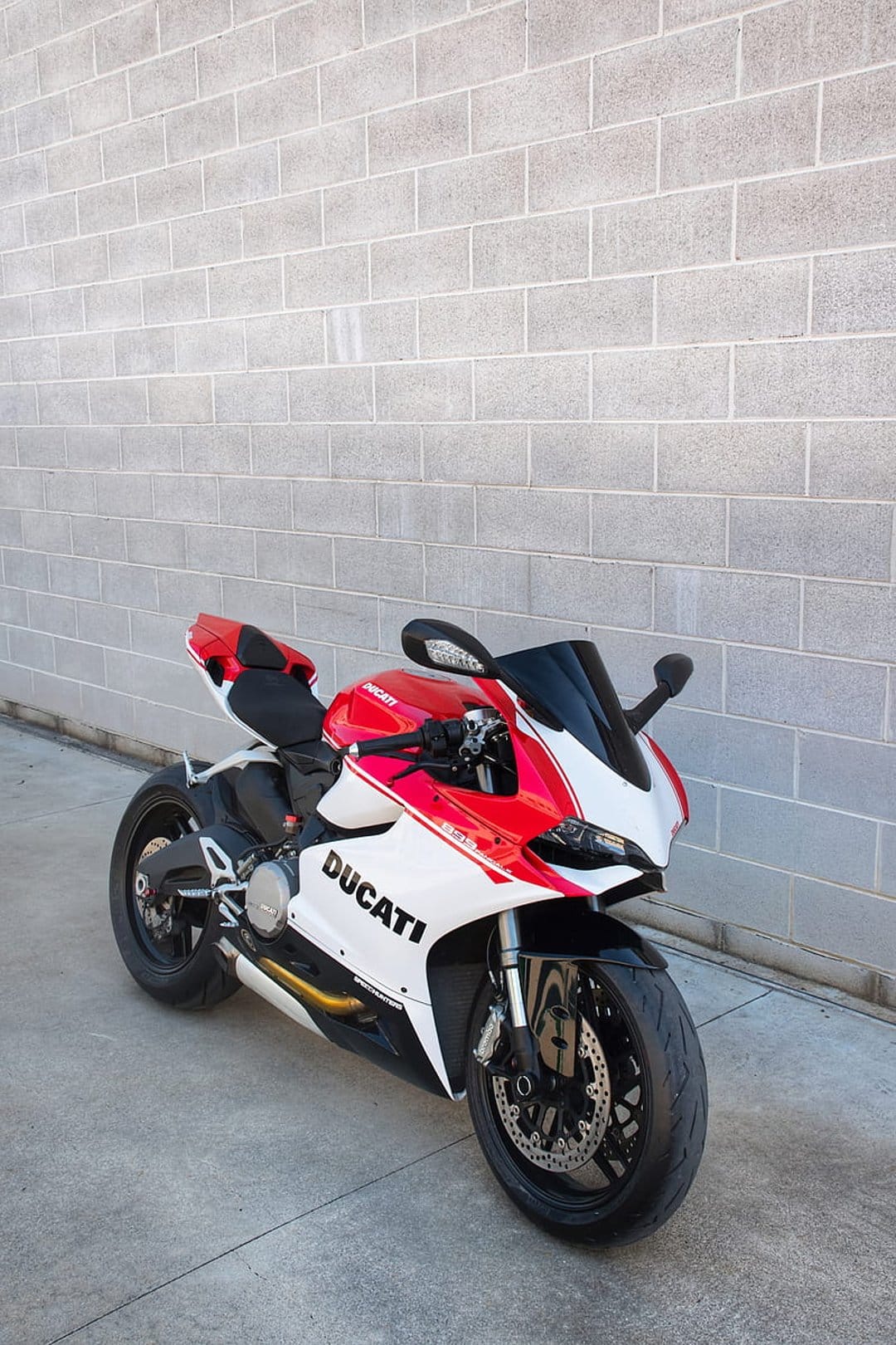 Red and white Ducati motorcycle parked on concrete against a light gray brick wall in a vertical portrait view.
