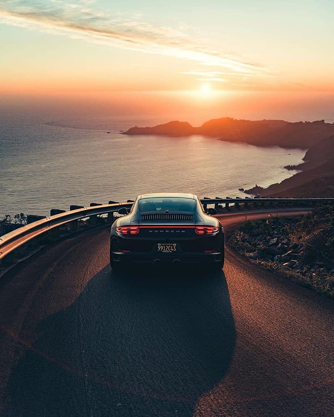 Black sports car driving on a coastal road during a golden sunset with ocean waves and cliffs in the background.