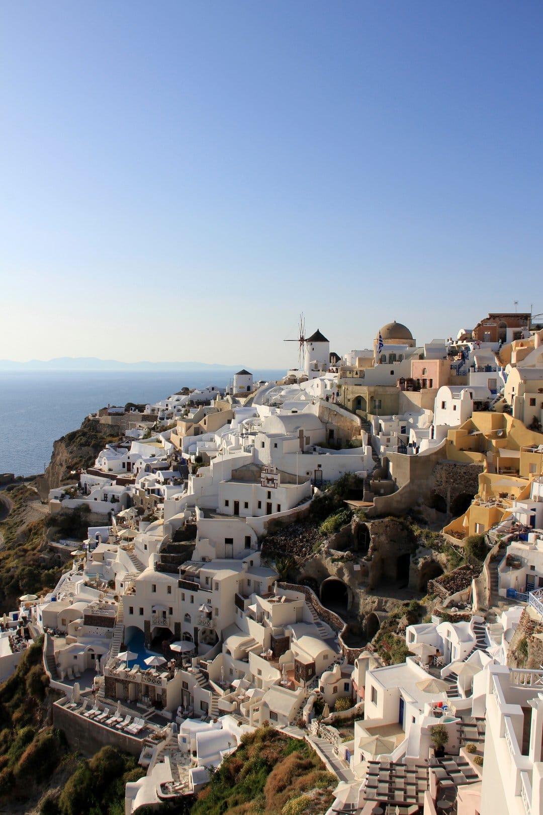 Whitewashed stone buildings with blue domes terrace down a steep volcanic cliff overlooking the Aegean Sea.